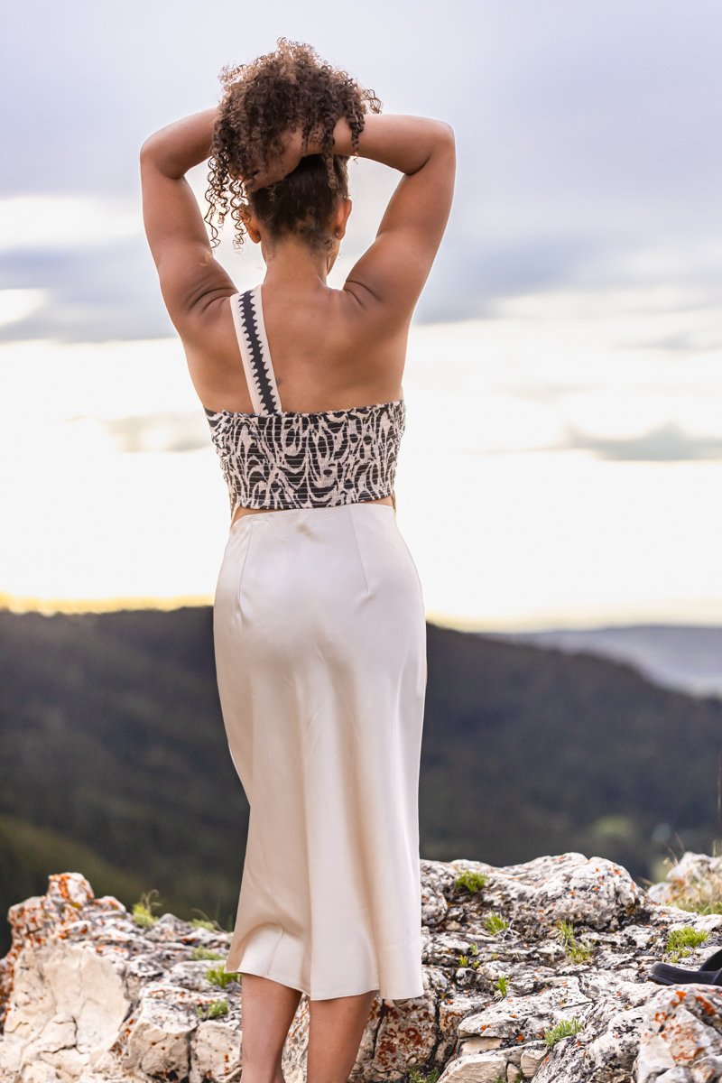 Woman on a mountain top in a pencil skirt, hair up, revealing her bare shoulders and back in a beautiful print blouse, creating a visual for her unique personal brand.