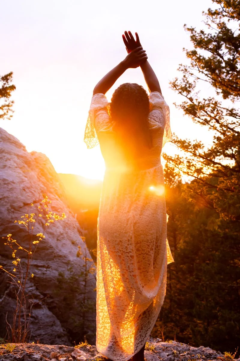 The sun glows around and through the white lace dress of a woman standing on a mountain top expressing her true self as part of her personal branding session near Bozeman Montana.