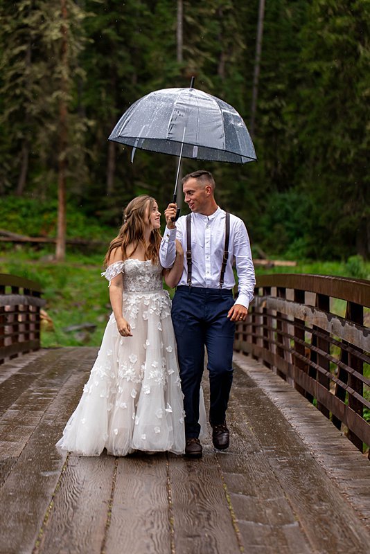 A bride and groom walking along a mountain path under an umbrella on a rainy day.