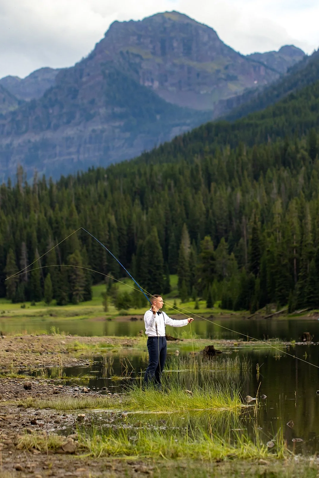 A groom in his wedding outfit, flyfishing below the looming mountains around Hyalite Reservoir.