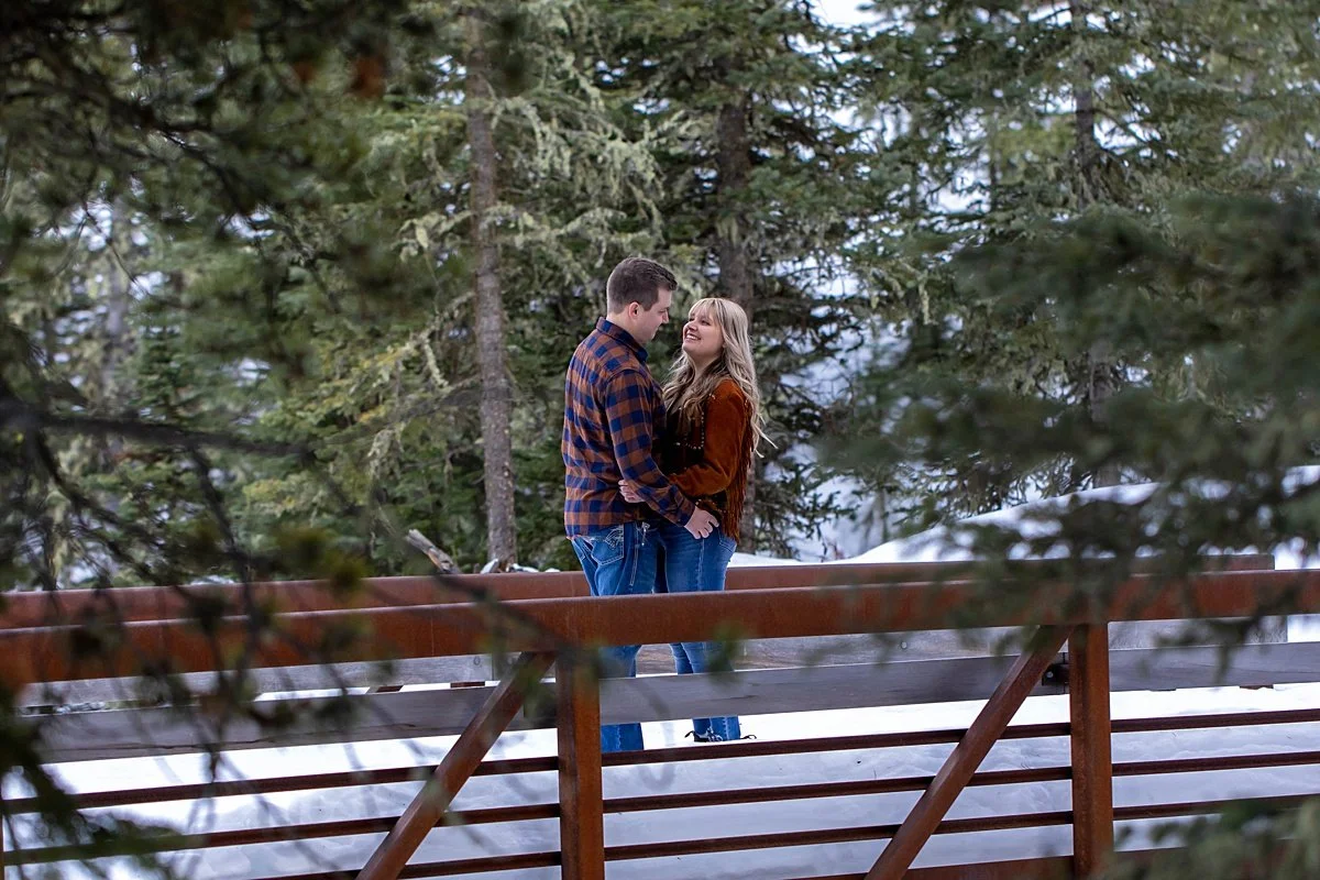 Couple on a bridge, engaging in happy eye framed by evergreens all around them.