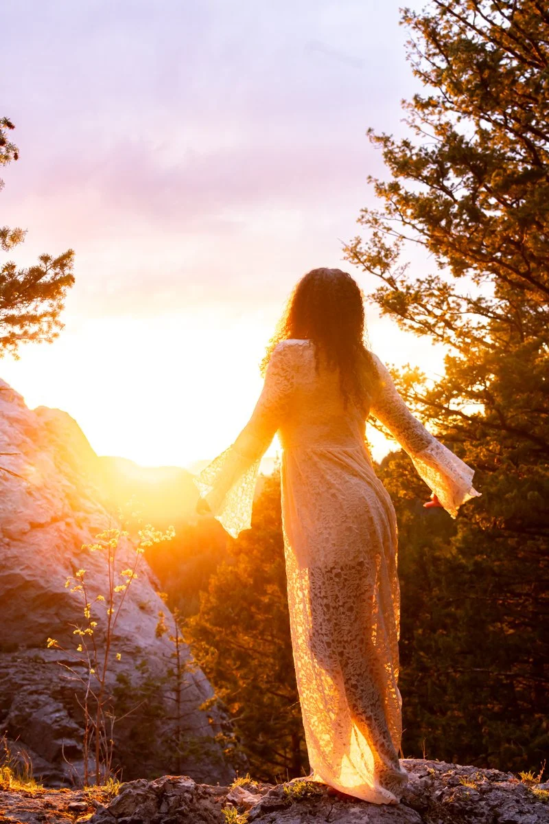 Woman standing in the trees, looking into a glowing sunset that is reflected in her white lace dress.