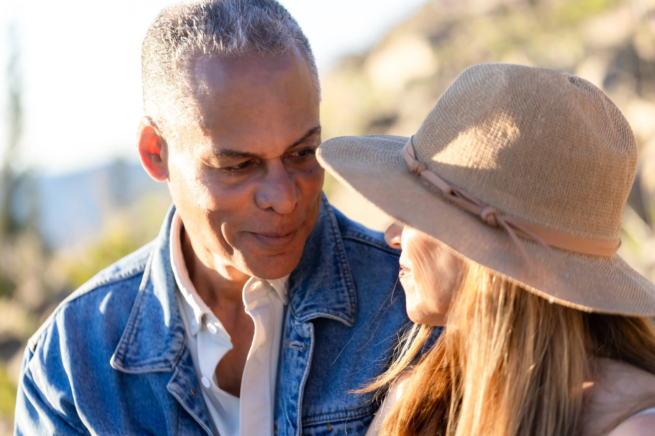 A mixed race couple huddled close together with the sun drenching them with warmth as they look at each other, both on the verge of bursting into laughter.