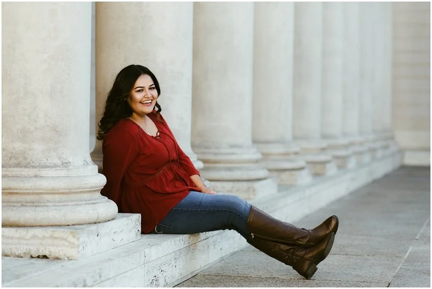 Senior girl sitting among the columns of a beautiful bit of architecture, her red shirt popping among the white structure, smiling, kicking back with her booted feet crossed in a relaxed and casual way.