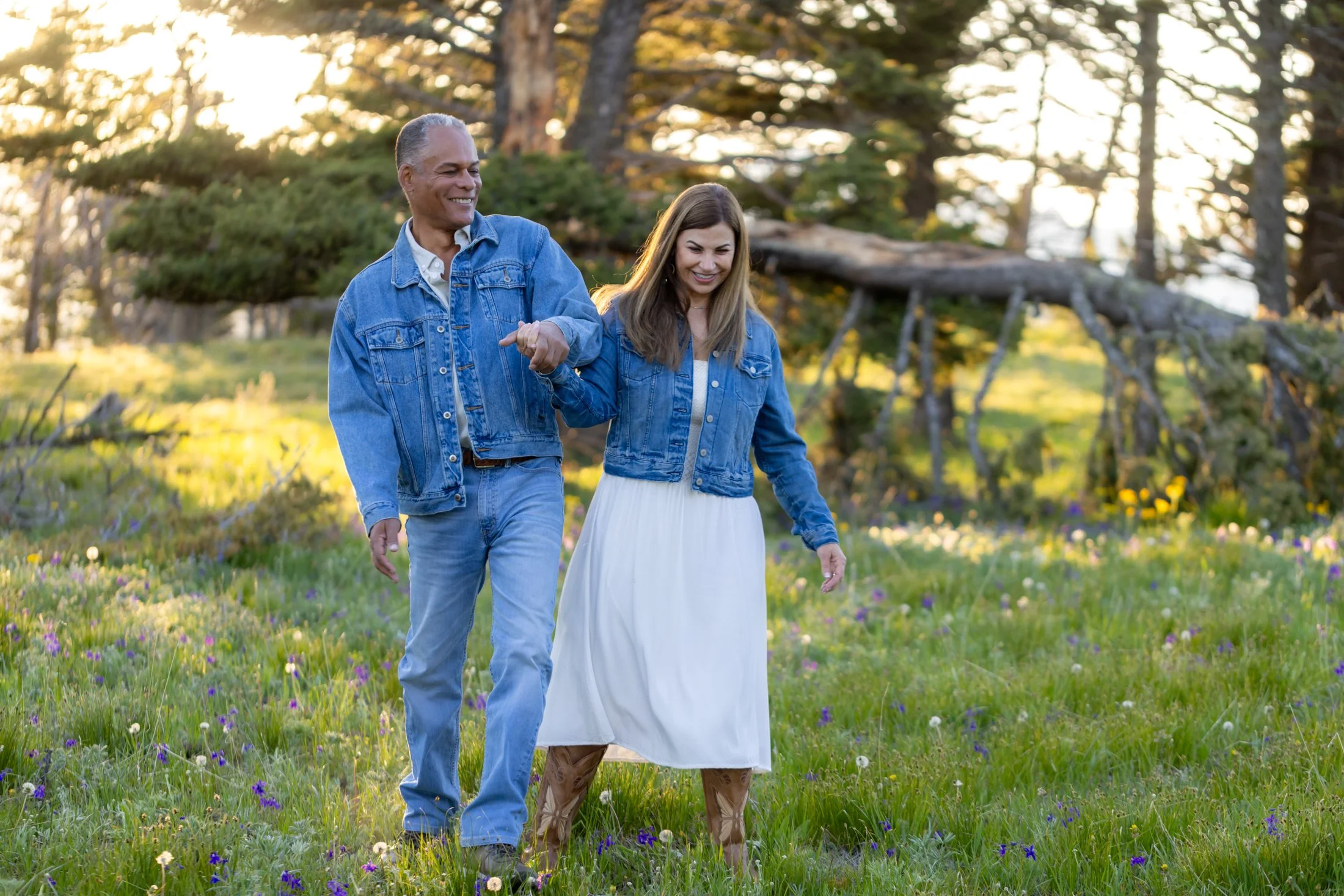An engaged couple playfully tapping hips together as they walk through a high mountain meadow during their Montana Engagement Sesion. Wildflowers and filtered sunlight surround their playful fun.