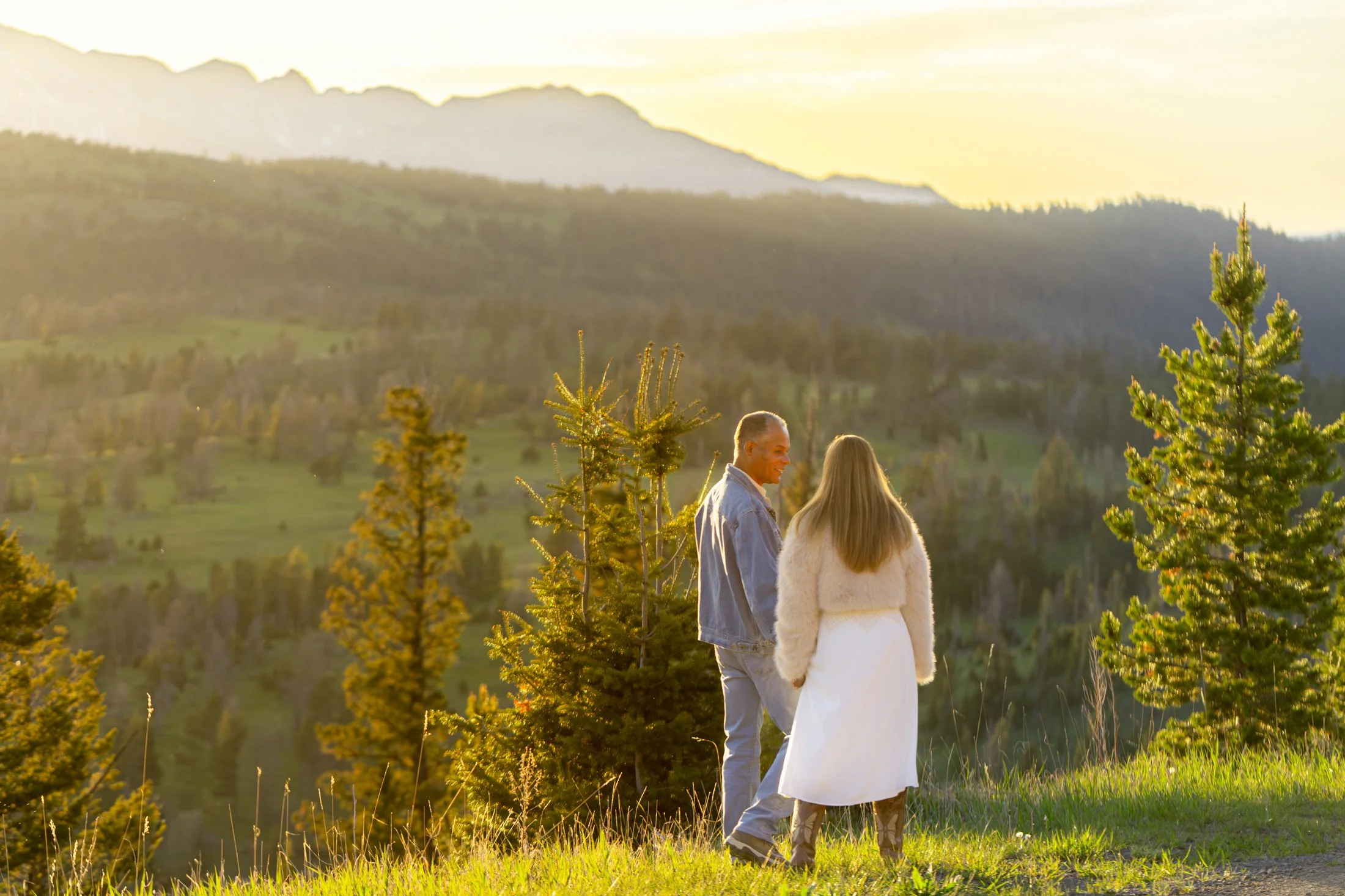 A couple walks along a beautiful  mountain overlook during their Montana Engagement Session. He looks at her with joy and connection as they stroll along the top of a grassy ridge.