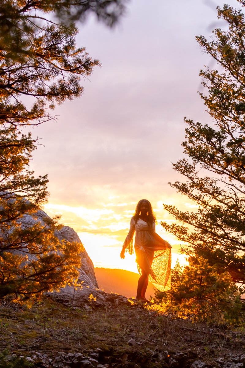 Woman backlit at sunset in the mountains, with the golden glow of the sky all around her and reflecting off of the rocks and trees that surround her in a mountain top setting.