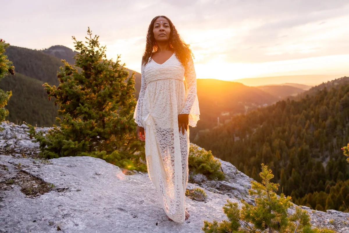 At sunset, on a mountain top, a beautiful woman with long curling hair stands in her power while her white lace dress captures the authentic femininity she brings to everything she does.