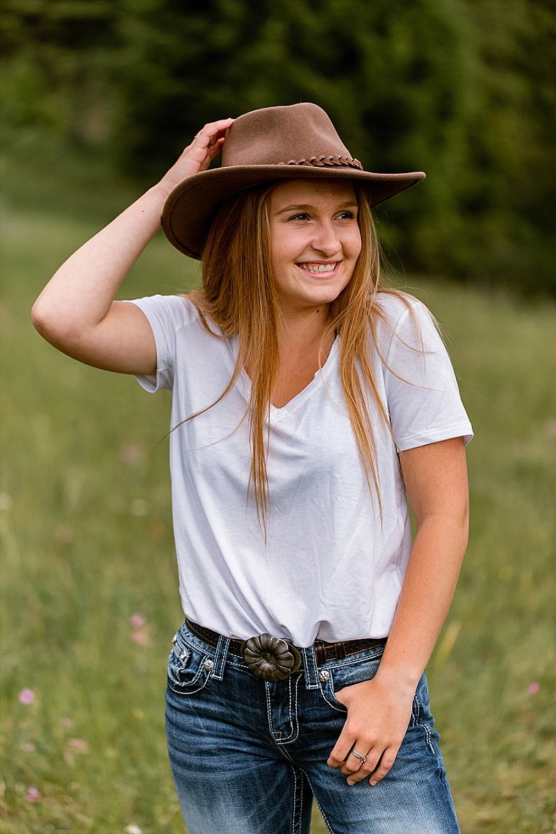 A high school senior girl in a mountain meadow with trees in the background. She is holding her hat and smiling big!
