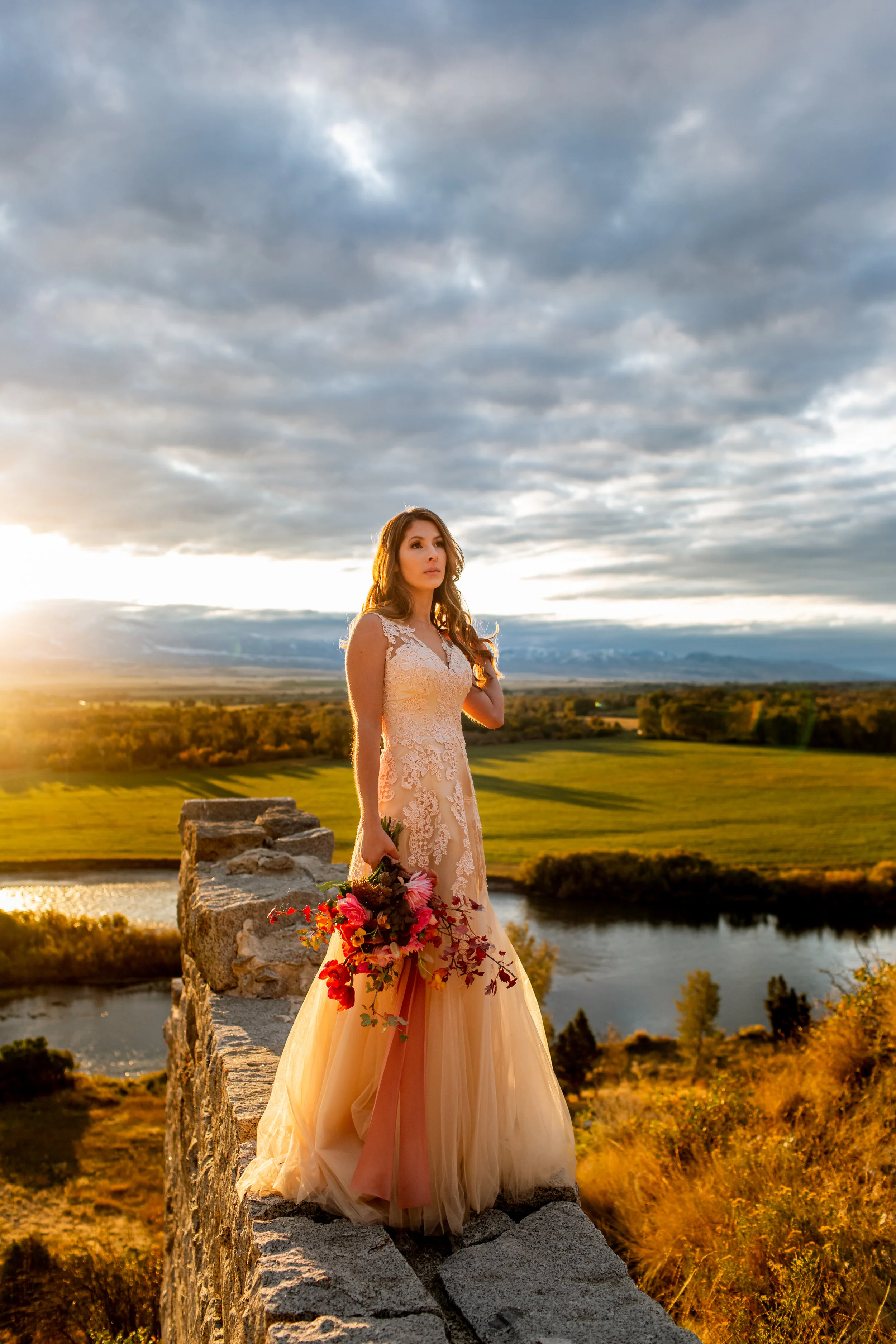 A bride standing on a stone wall that overlooks the Jefferson River in Whitehall Montana. Storm clouds float above her, and all the colors of the landscape are glowing with sunset's golden light.