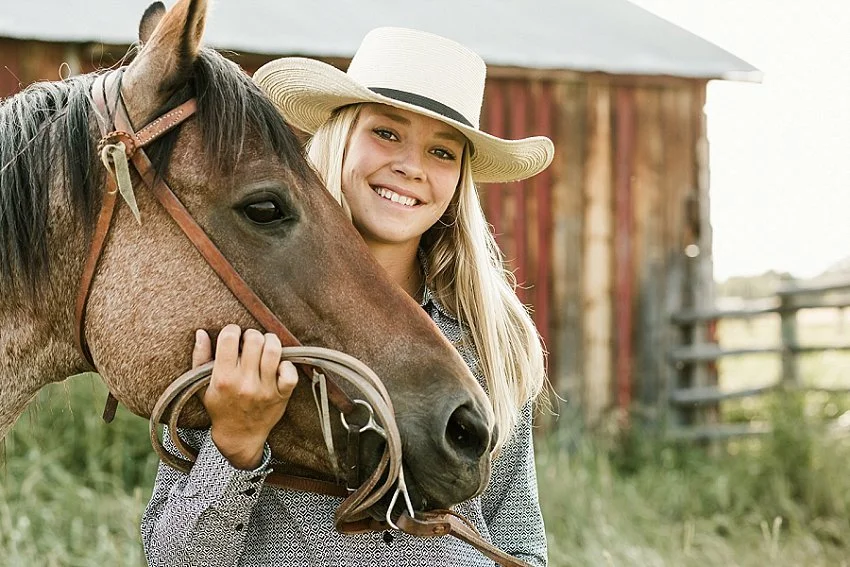 A High School Senior from the Bozeman Montana area with her horse, in front of an old red outbuilding surrounded by grass and fence.