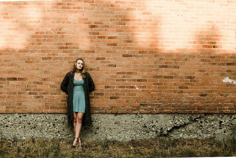 A senior girl up against a brick wall in Bozeman Montana, with the sun hitting the building as she is tucked into the shade of the lower half of the image.