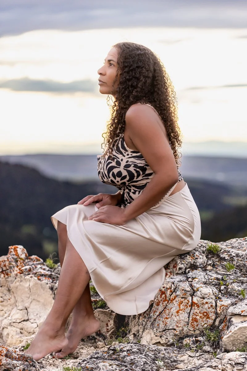 Woman dressed professionally in a silk skirt and print blouse, sitting high on a mountain top with a beautiful sunset behind her - showing her personal brand is connected to the environment where she works.