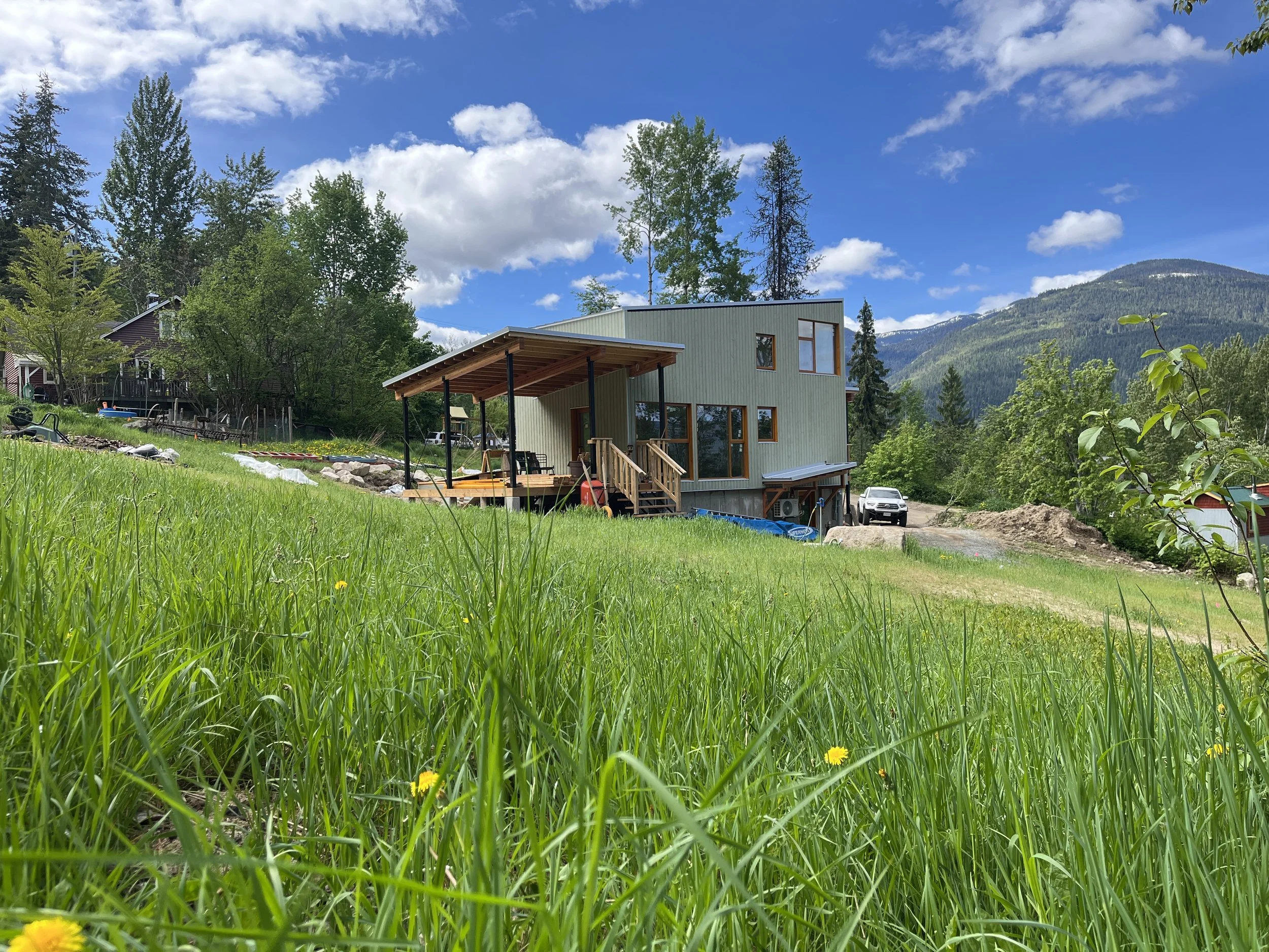 Modern house with a wooden deck and stained glass windows, surrounded by green grass and trees, with mountains in the background under a partly cloudy sky.