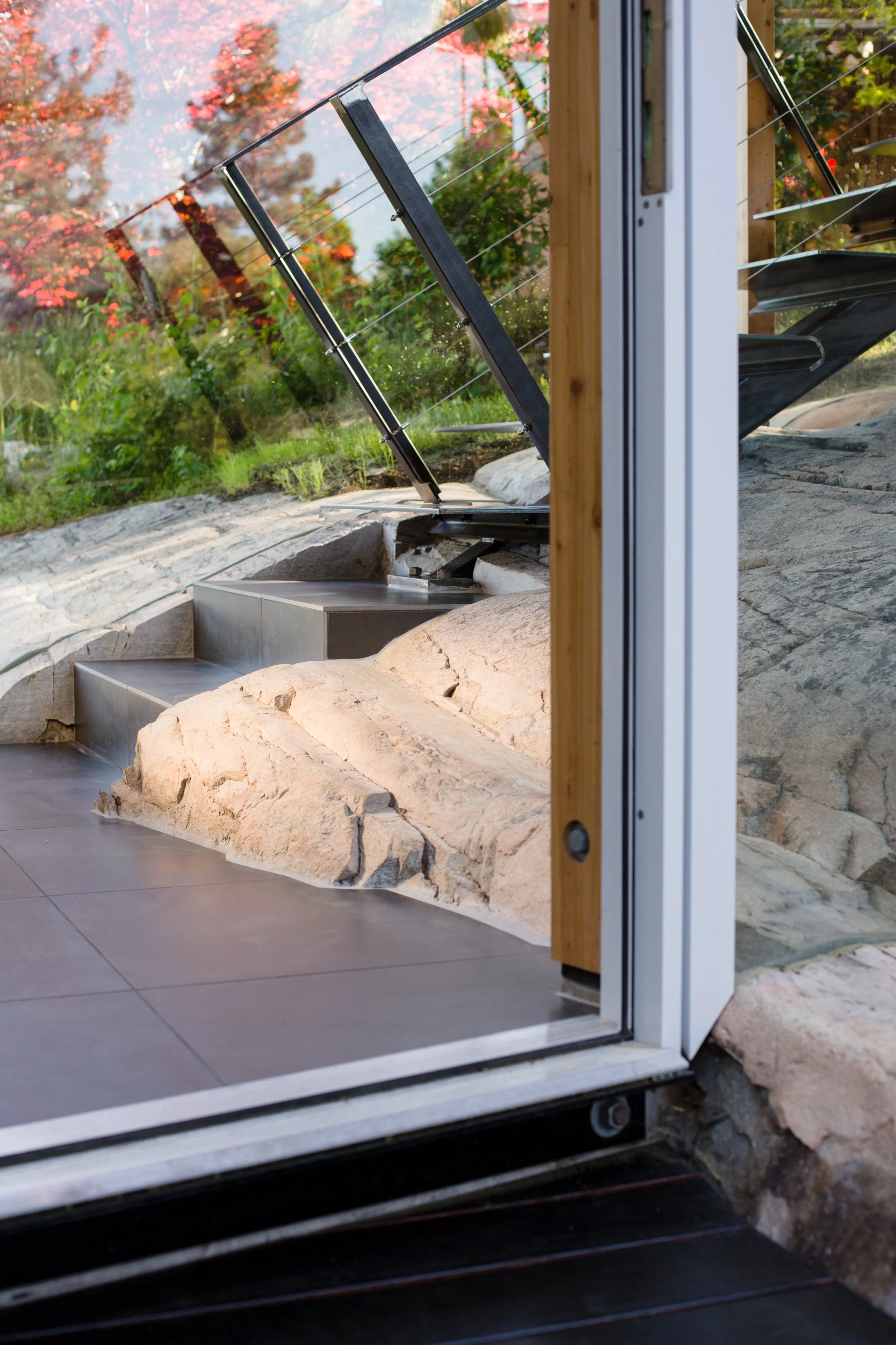 View through a glass door showing stone steps, rocks, trees with fall foliage, and lush greenery outside.