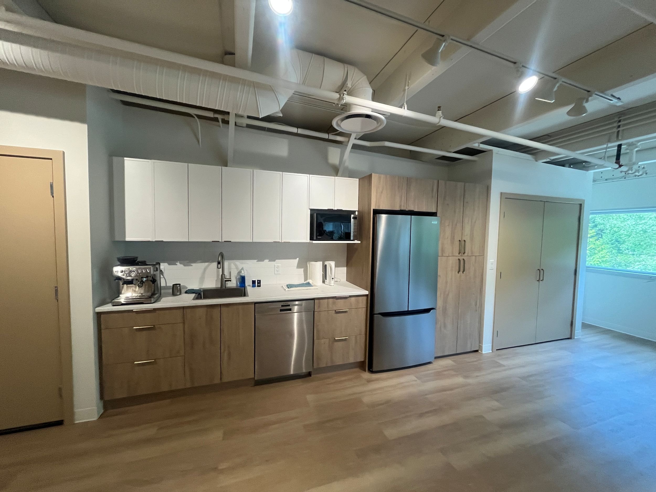 Modern kitchen with white and wood cabinets, stainless steel refrigerator, microwave, and coffee machine. Open space near large window with sliding door, wood flooring, and visible ceiling pipes and lights.
