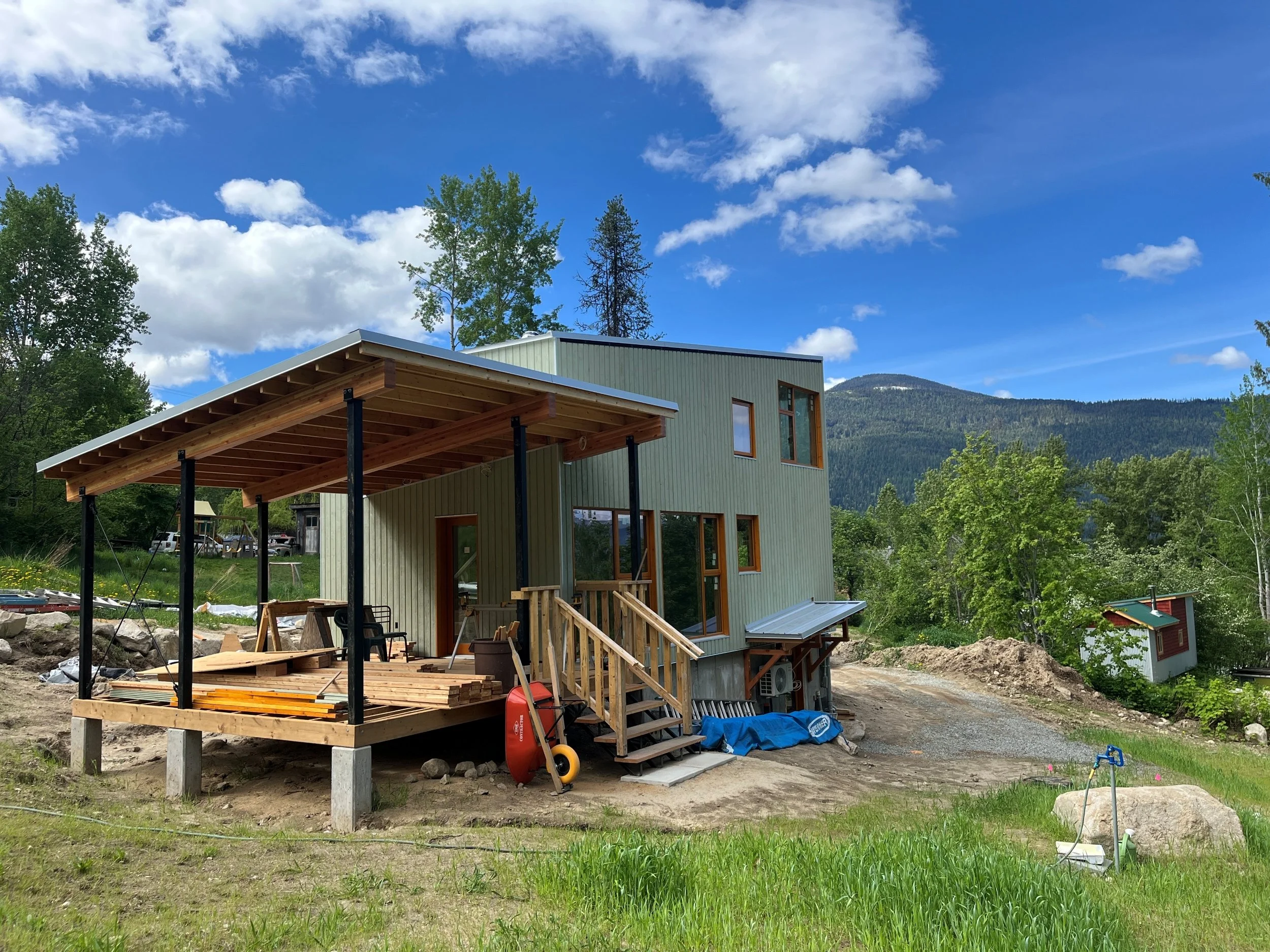 Newly built modern house with a wooden deck under construction, situated in a green landscape with mountains in the background, blue sky, and scattered clouds.