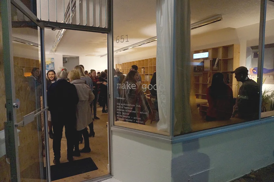 Group of people gathering inside a modern art gallery or exhibition space, seen from the outside through large glass windows, with some standing and some sitting, engaged in conversation and viewing art displays.