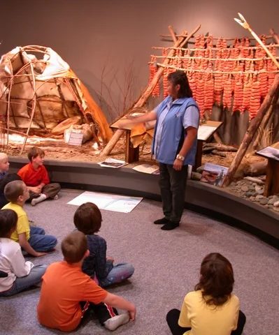 A woman standing and speaking to a group of children sitting on the floor in a museum exhibit about indigenous culture, with traditional artifacts and a imitation dwelling in the background.