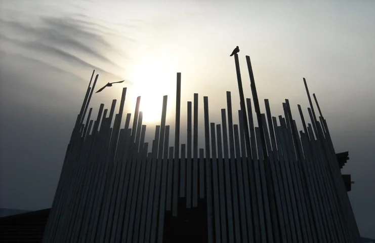 A modern wooden structure with vertical slats forming a circular shape, with the sun setting behind it and a bird perched on one of the slats.