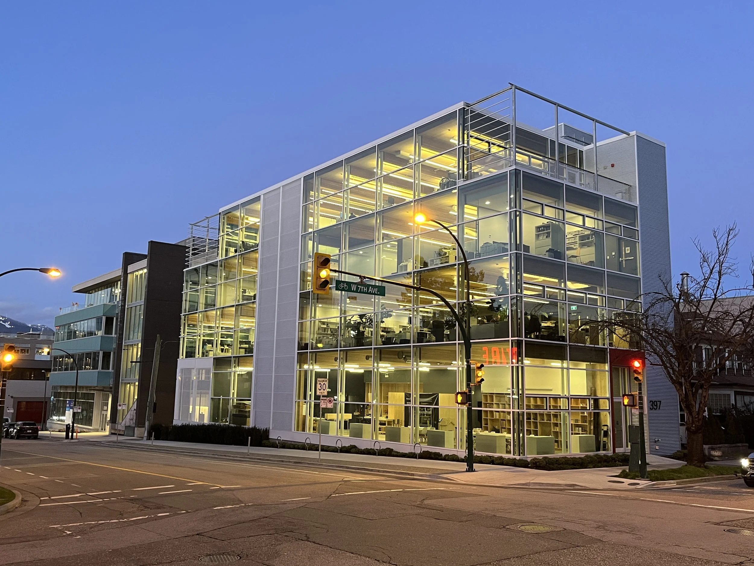 Modern glass building with interior lights on, located at a street corner with traffic signals and street signs, and surrounded by parked cars and leafless trees at dusk.