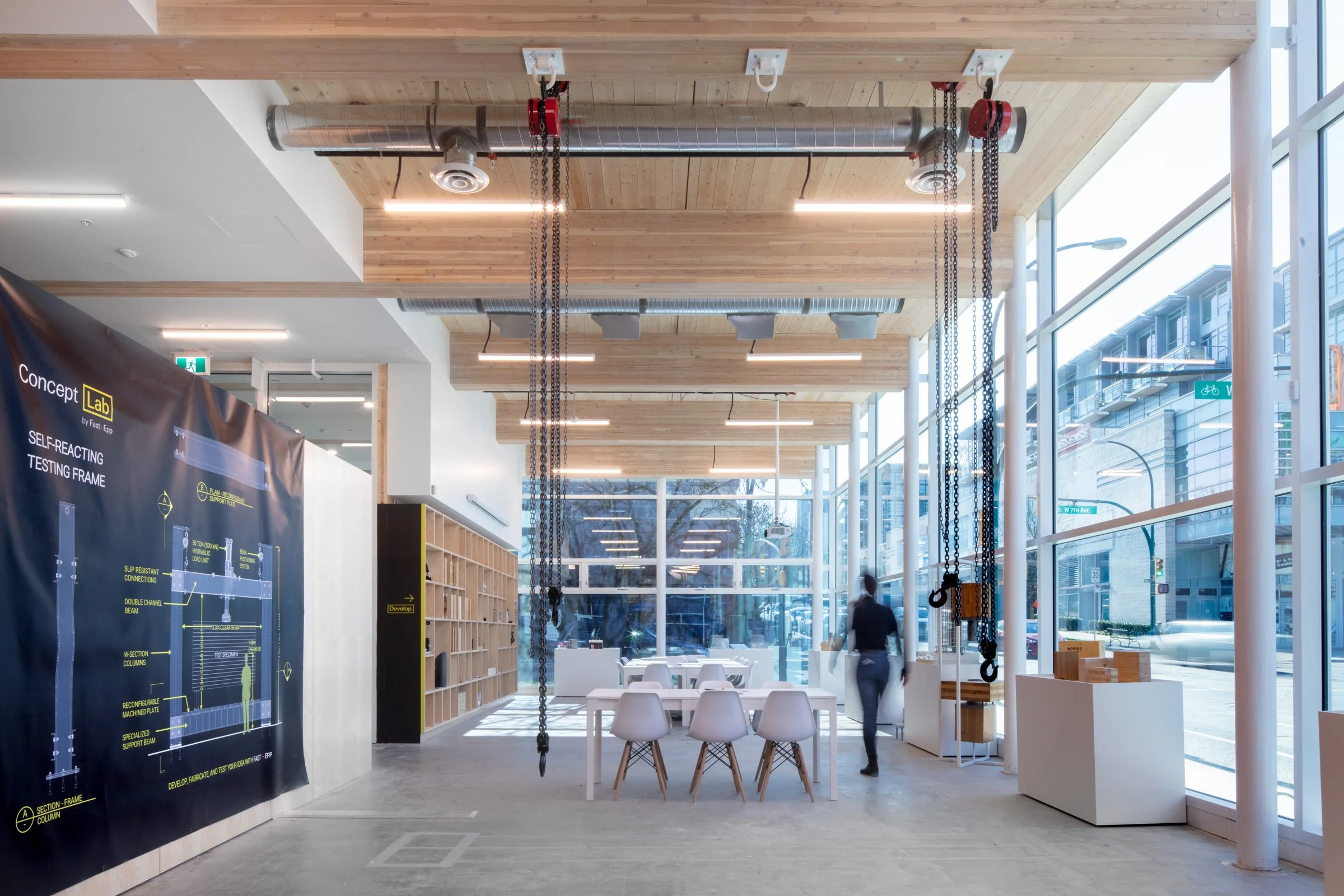 Modern interior of a bright, airy coworking space with large floor-to-ceiling windows, wooden ceiling panels, hanging chains from lift equipment, tables and chairs, and a large technical poster on the wall.