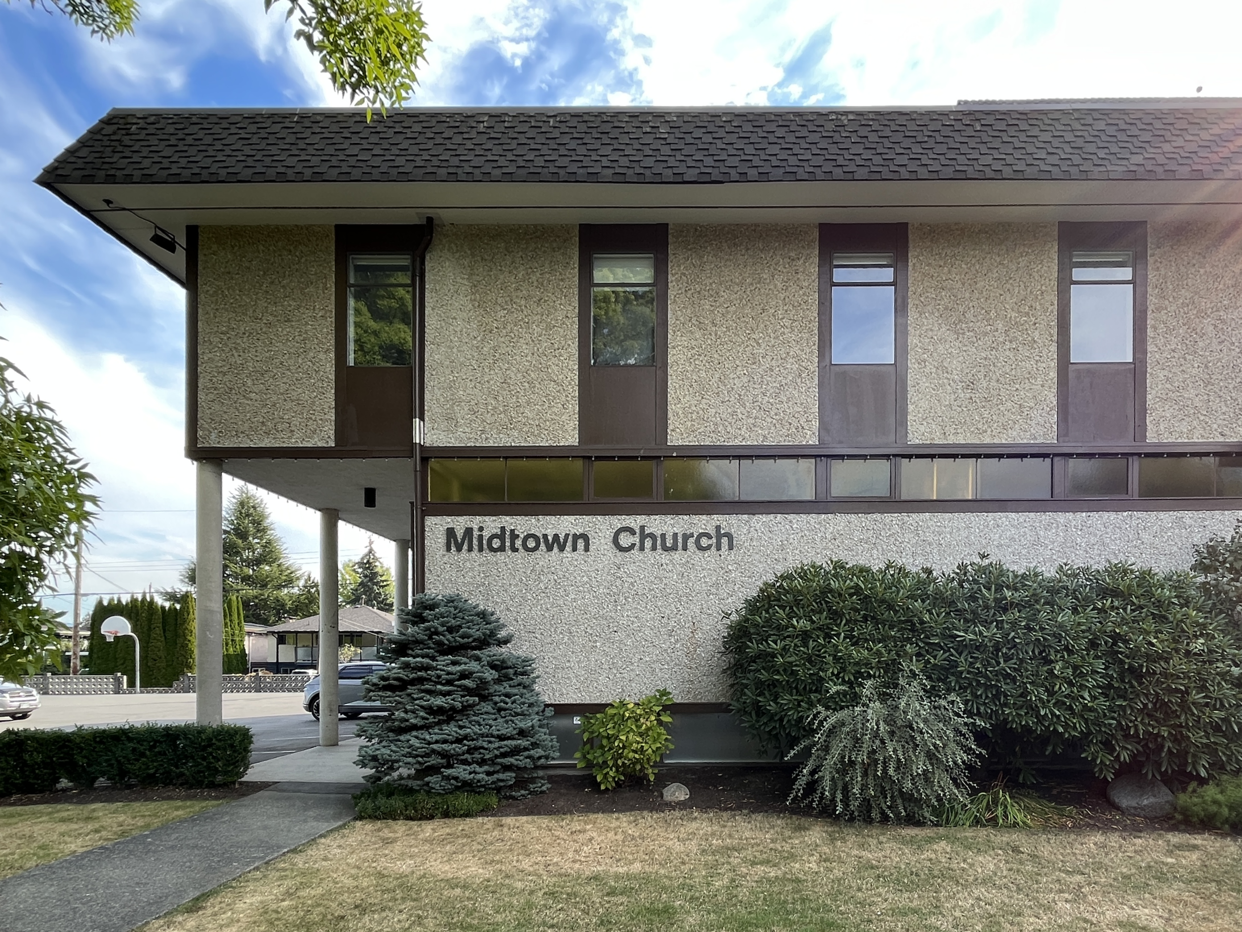 Exterior view of the pre-renovation Midtown Church building surrounded by trees and shrubbery, with a car parked nearby and the church's name on the wall.