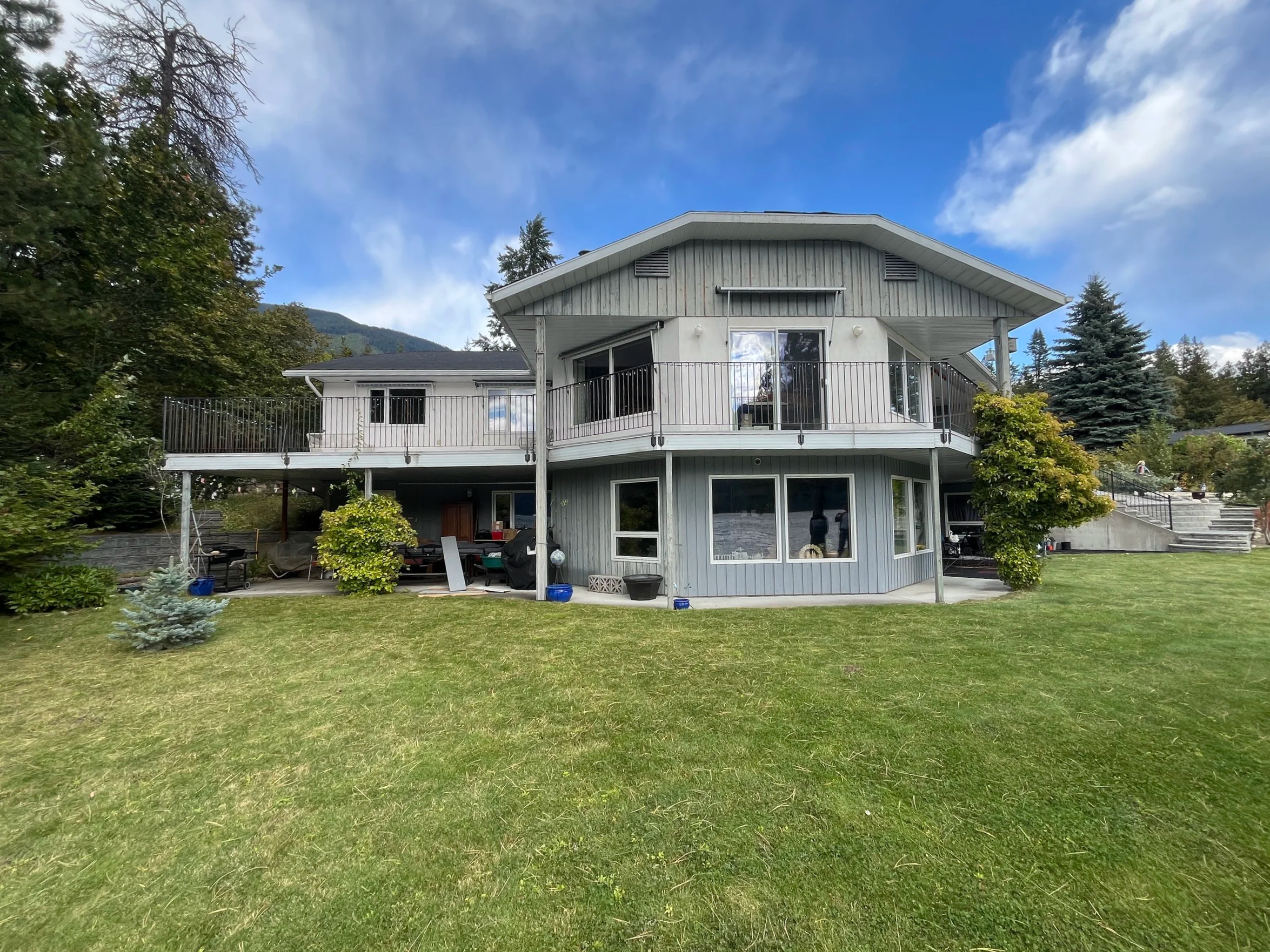 A two-story house with a large balcony on the upper level, surrounded by a green lawn and trees, with a partly cloudy sky above.