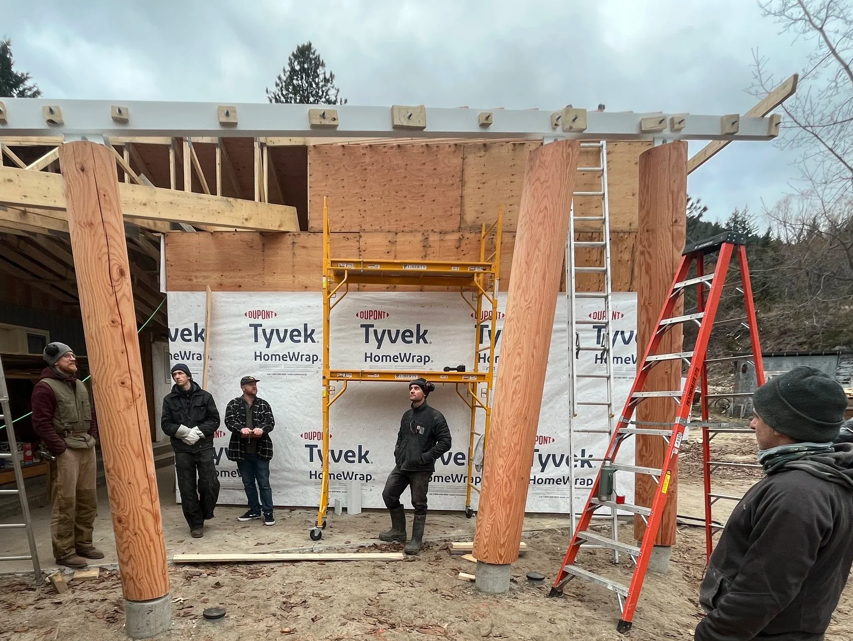 Group of people at a construction site with wooden beams, scaffolding, and ladders, building a structure.