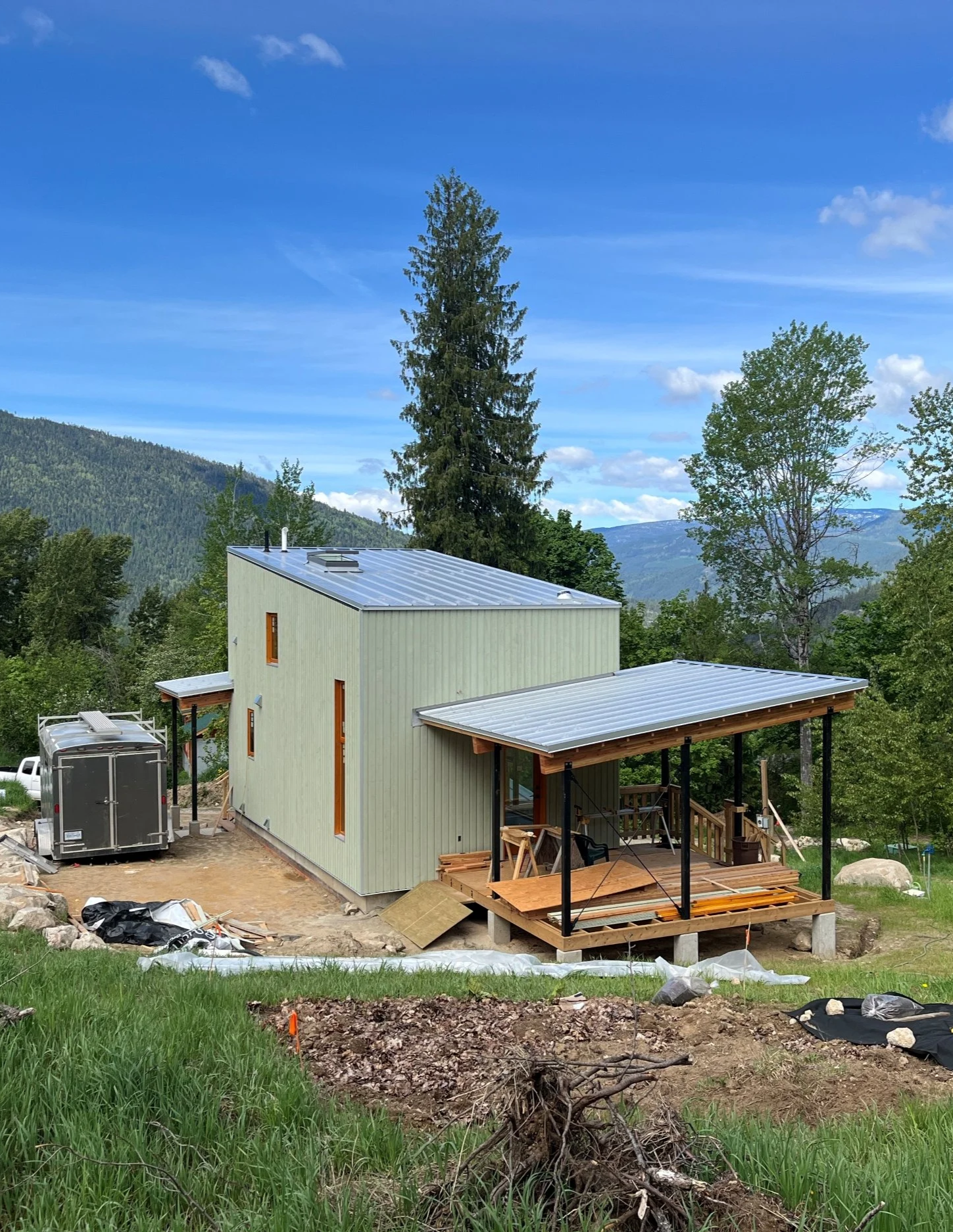 A house under construction on a hillside with green trees and mountains in the background, blue sky with white clouds.