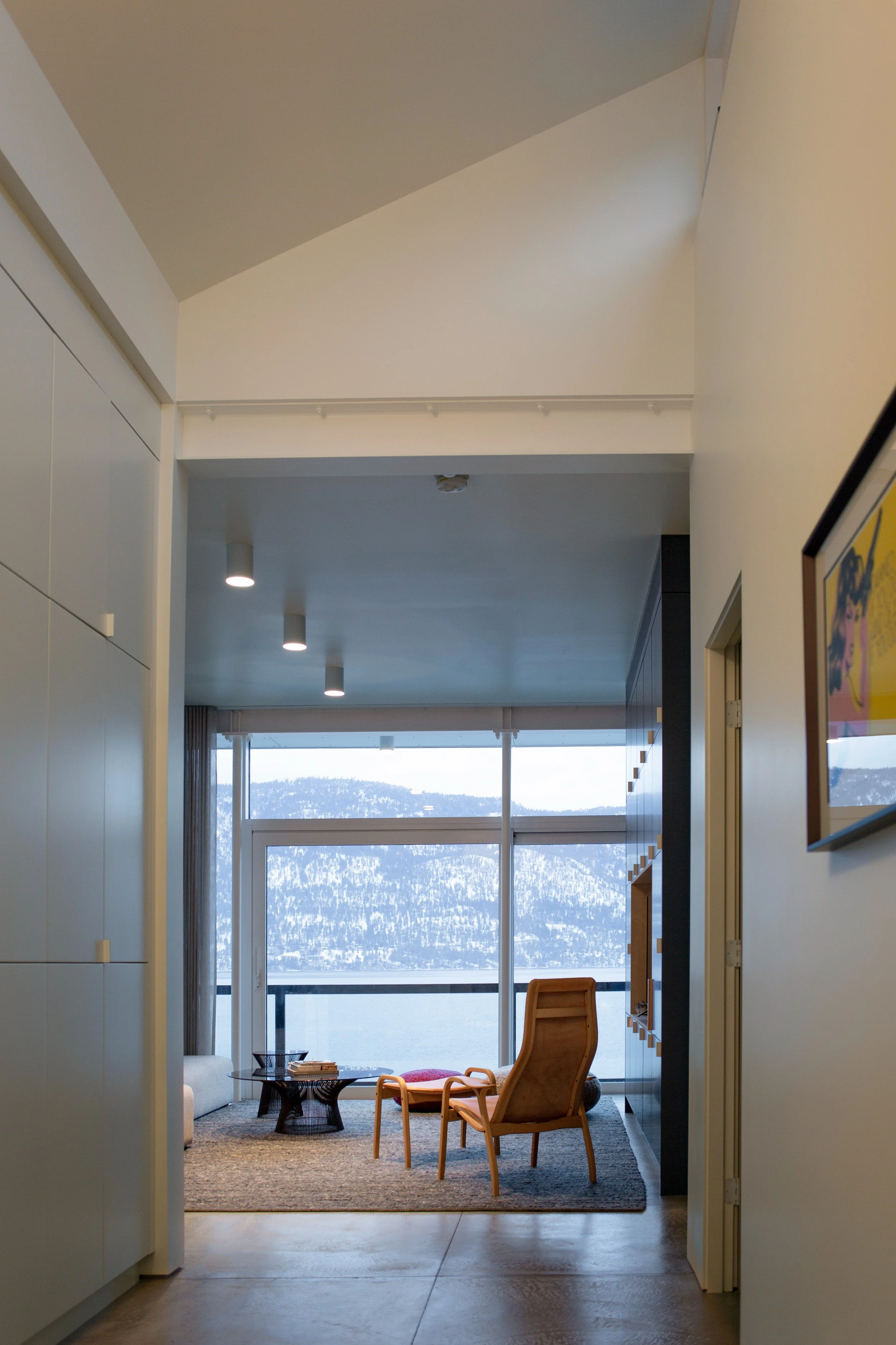 Interior view of a modern room with large windows showing snow-covered mountains outside, featuring lounge chairs, a rug, a small table, and wall decorations.