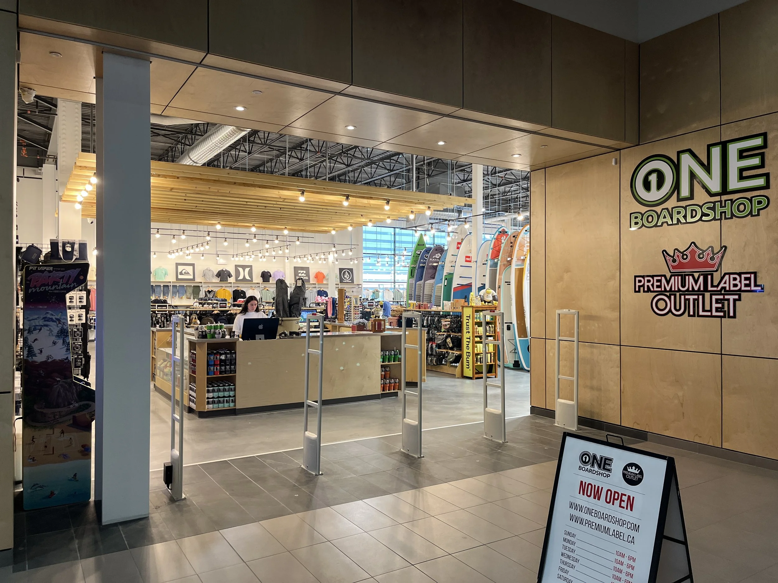 Interior view of a retail store called One Boardshop, displaying surfboards, apparel, and accessories, with a sign indicating it is now open.