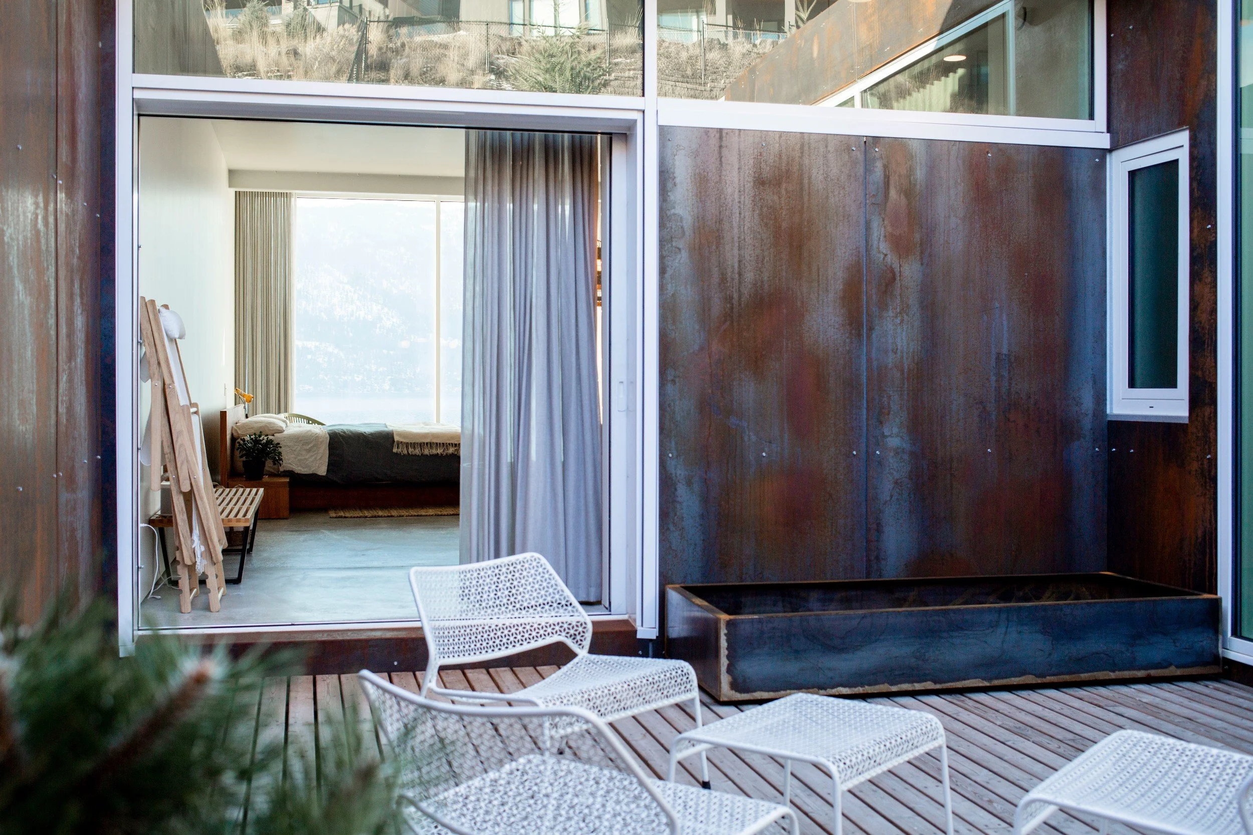 A modern indoor-outdoor living space featuring a wooden deck with white chairs, a large planter, and a sliding glass door leading to a bedroom with large windows, curtains, and simple furniture.