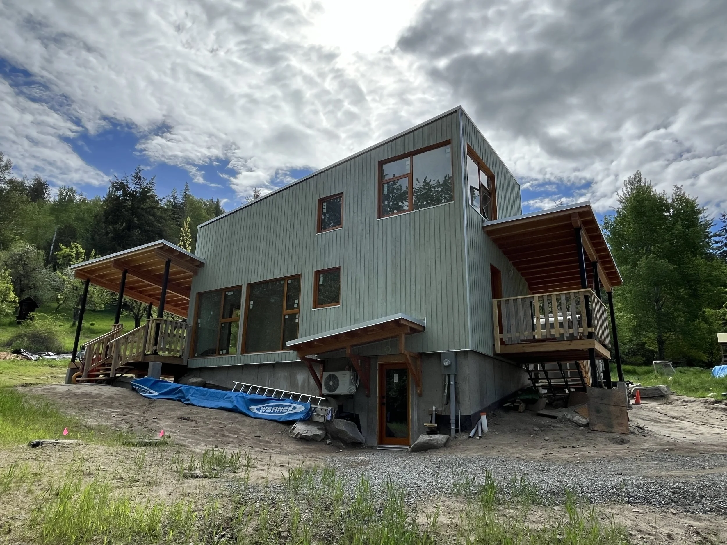 Newly constructed multi-story house with green exterior walls, large windows, and wooden balconies, set on a dirt lot with construction equipment and surrounding trees under a partly cloudy sky.