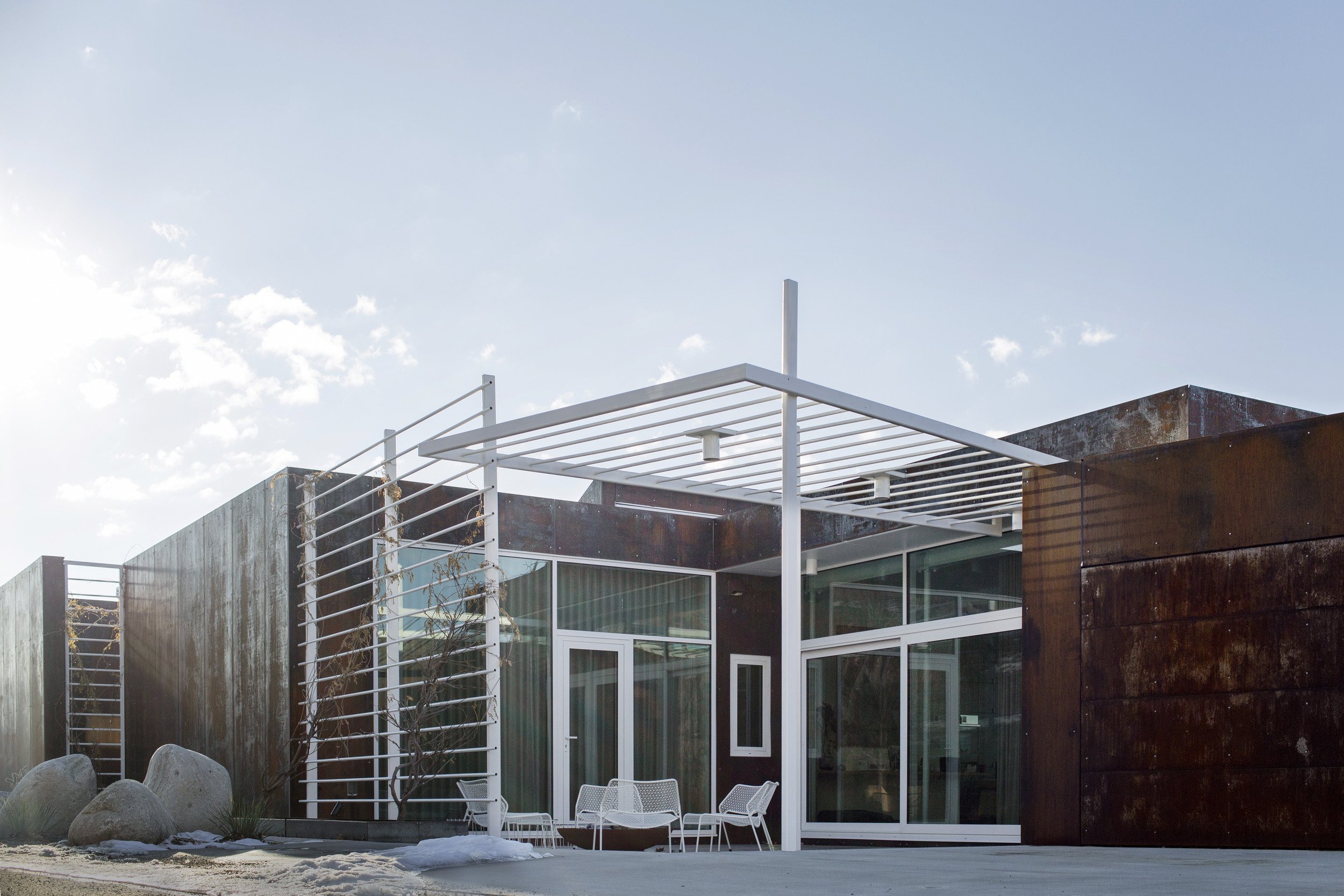 Modern house with rust-colored exterior walls, large glass windows and doors, and a white metal pergola on a sunny day.