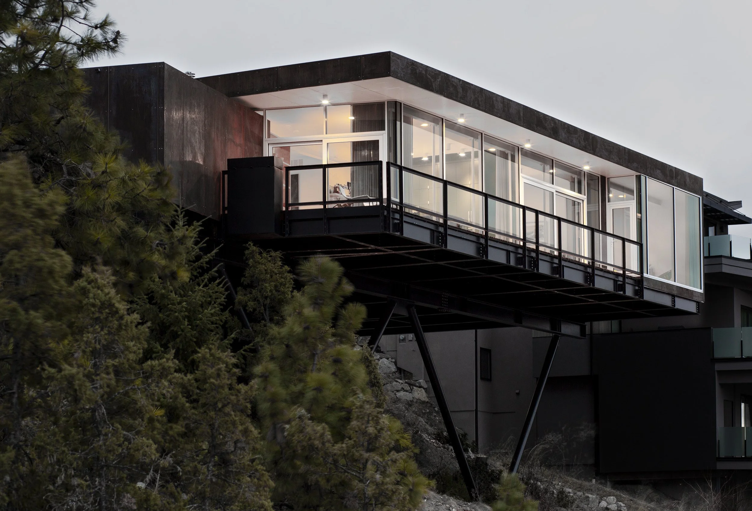 Modern house cantilevered over a cliff with large glass windows, metal railing, and supported by V-shaped stilts, surrounded by trees.