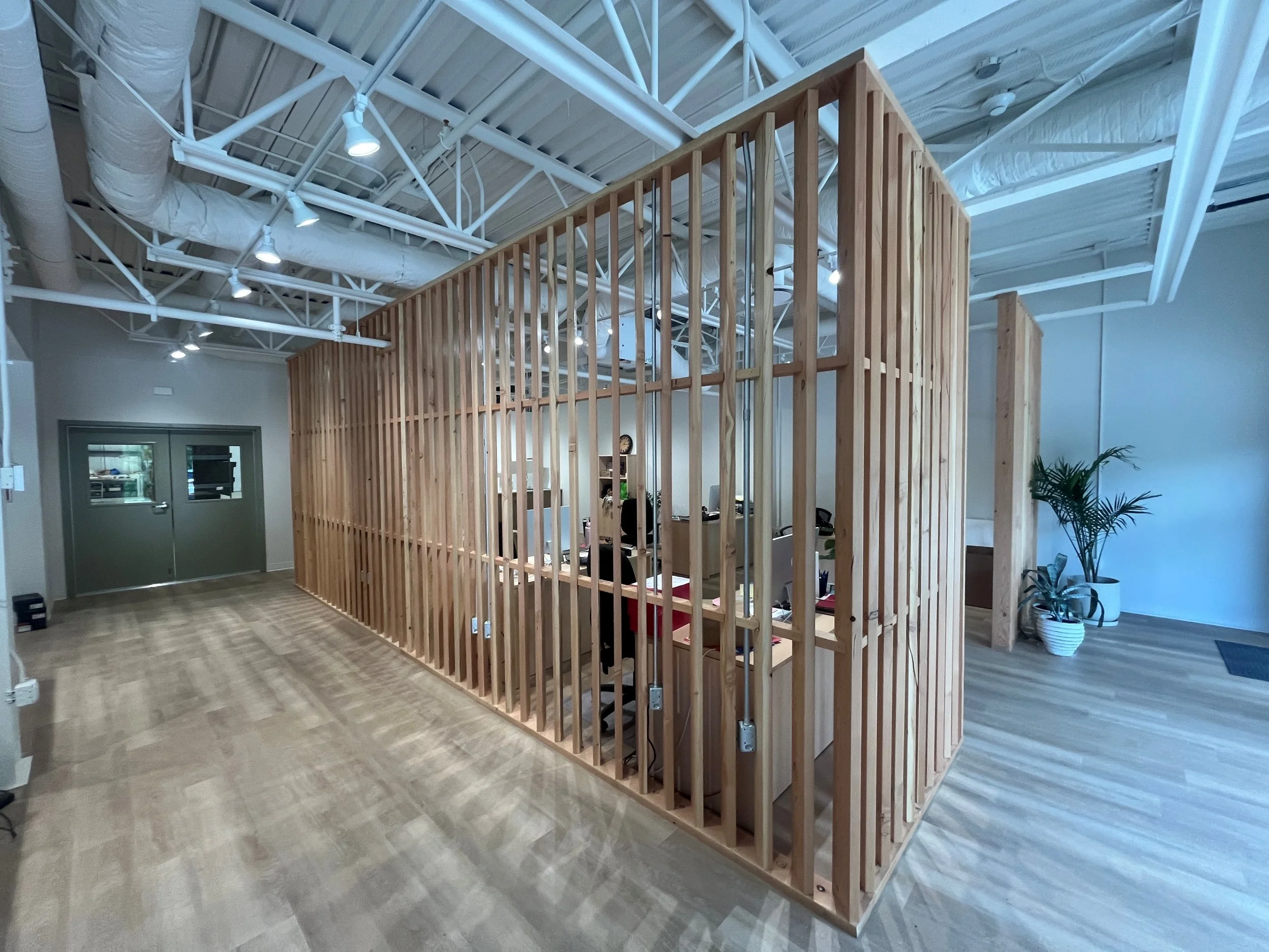 An office space with a wooden cubicle wall and an industrial ceiling with exposed pipes and ductwork, illuminated by multiple ceiling lights.