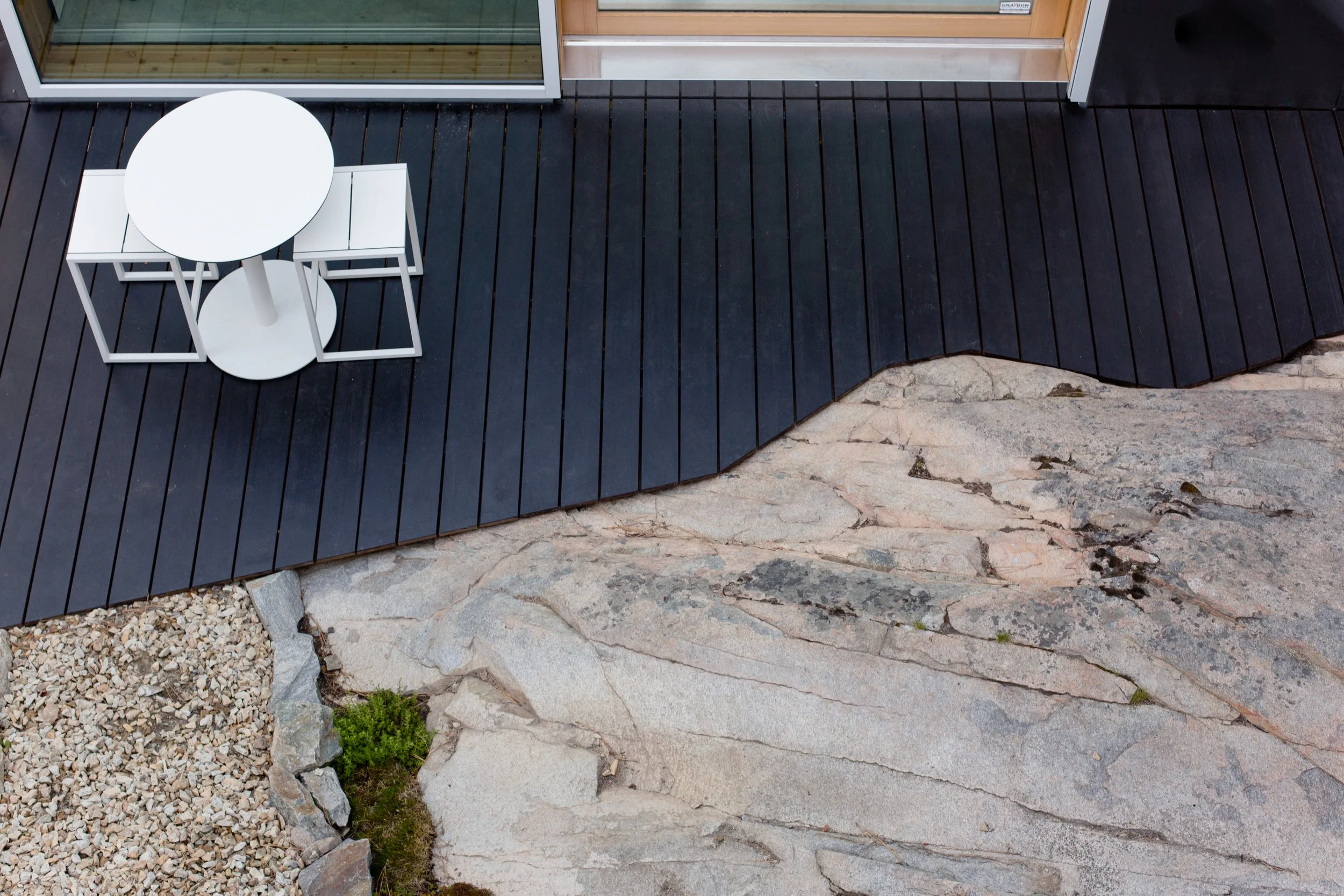 An outdoor patio with black wooden decking, a white round table, and matching white chairs, adjacent to a stone surface with small rocks and patches of grass.