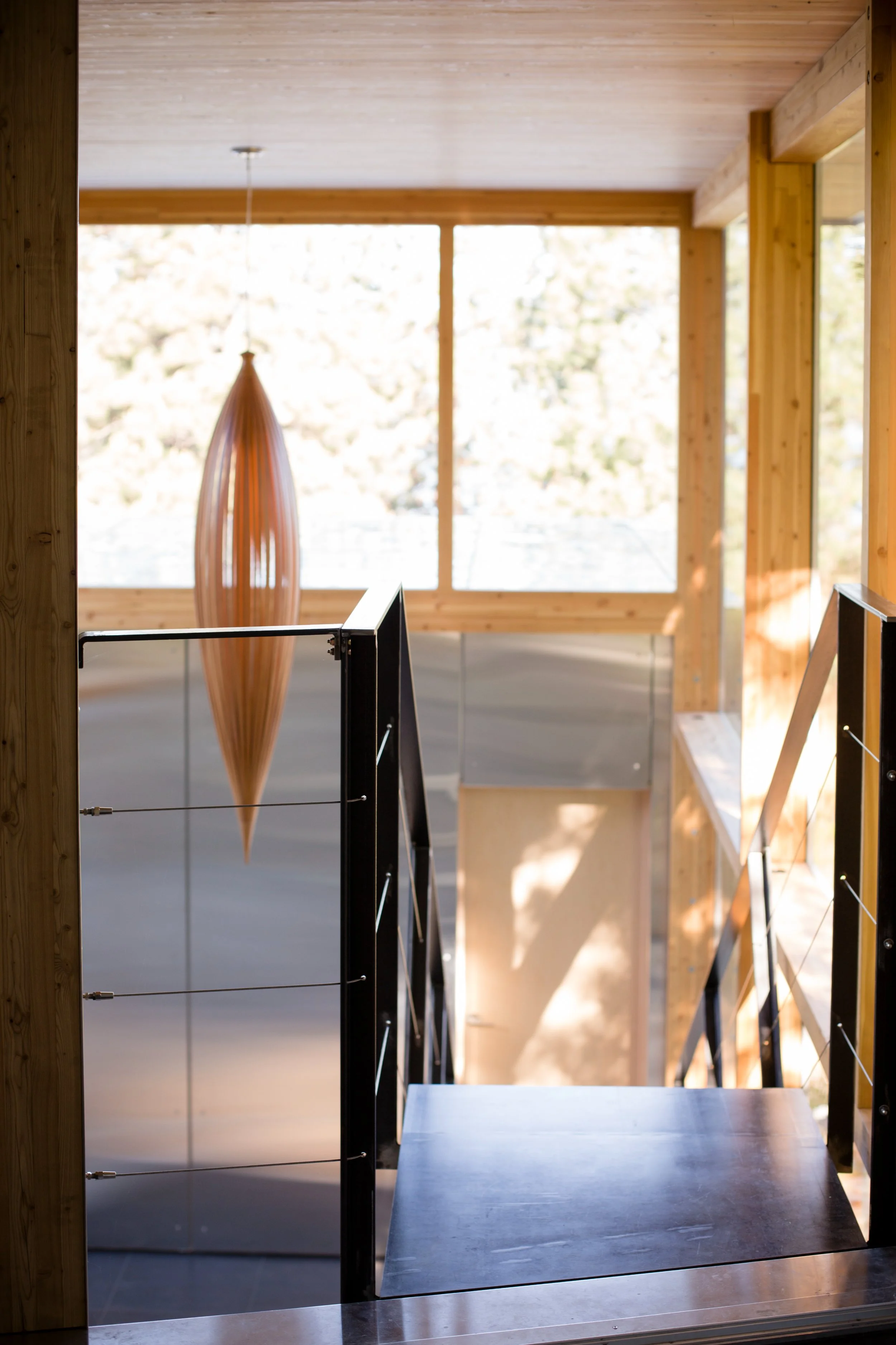 Wooden staircase with black metal railings leading to a sunlit room with large windows and a wooden ceiling, featuring a tall, elongated wooden pendant light.