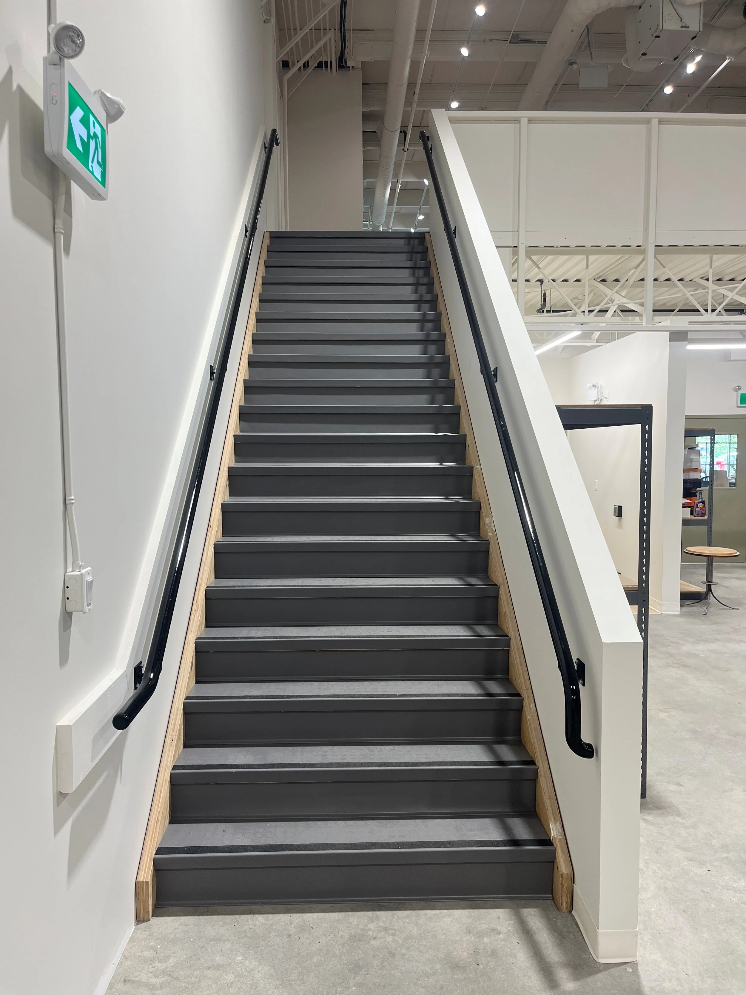 Indoor staircase with black treads and wooden side panels, black handrails on both sides, in a modern, well-lit space with white walls and ceiling pipes. An emergency exit sign is on the wall to the left.