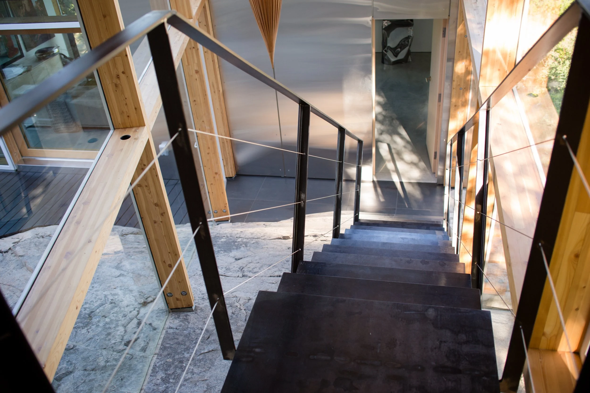 Interior view of a modern staircase with black steps, metal handrails, and wooden walls, leading down to a tiled floor and glass-walled rooms with reflections and natural lighting.