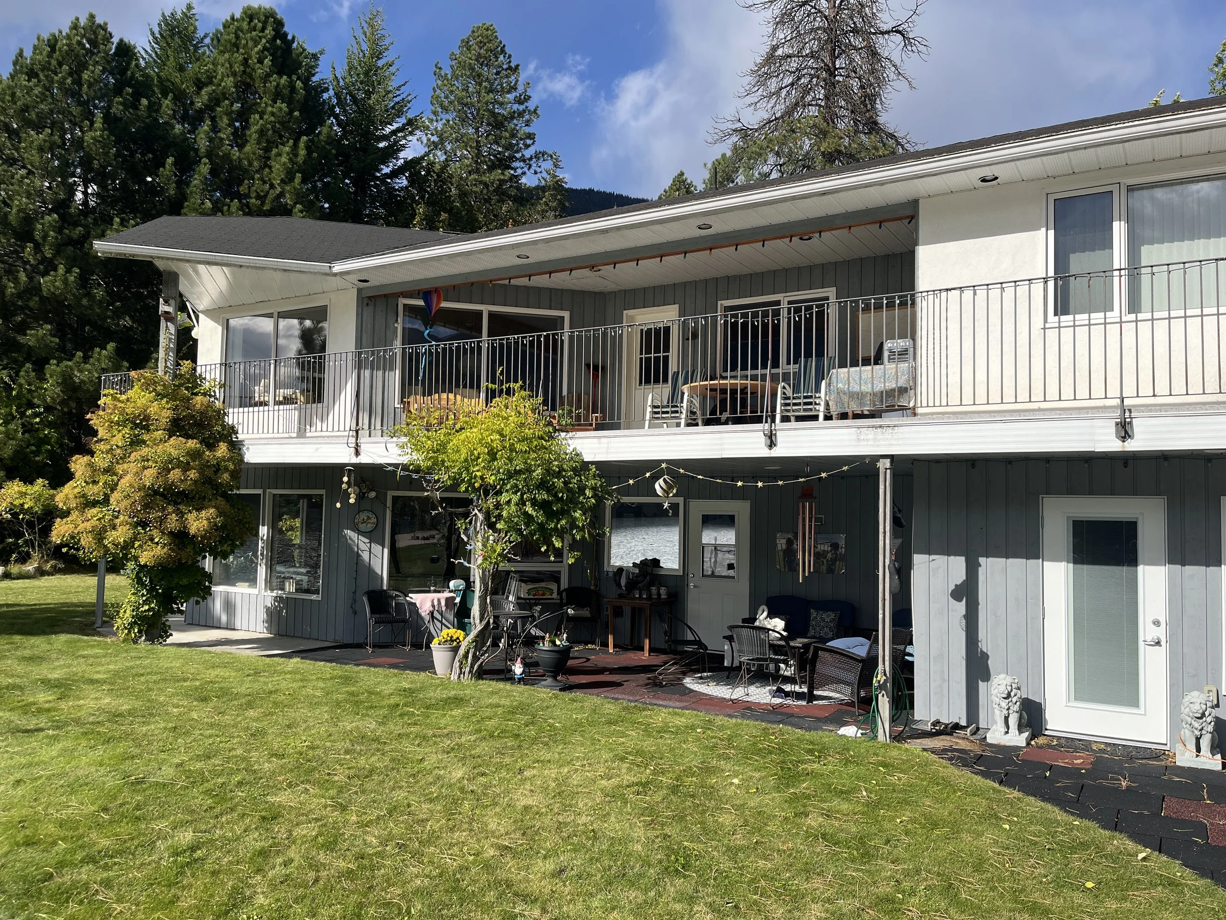 A two-story house with an upper balcony and lower patio, surrounded by a grassy yard with trees, decorated with plants, outdoor furniture, and ornaments in a sunny setting.
