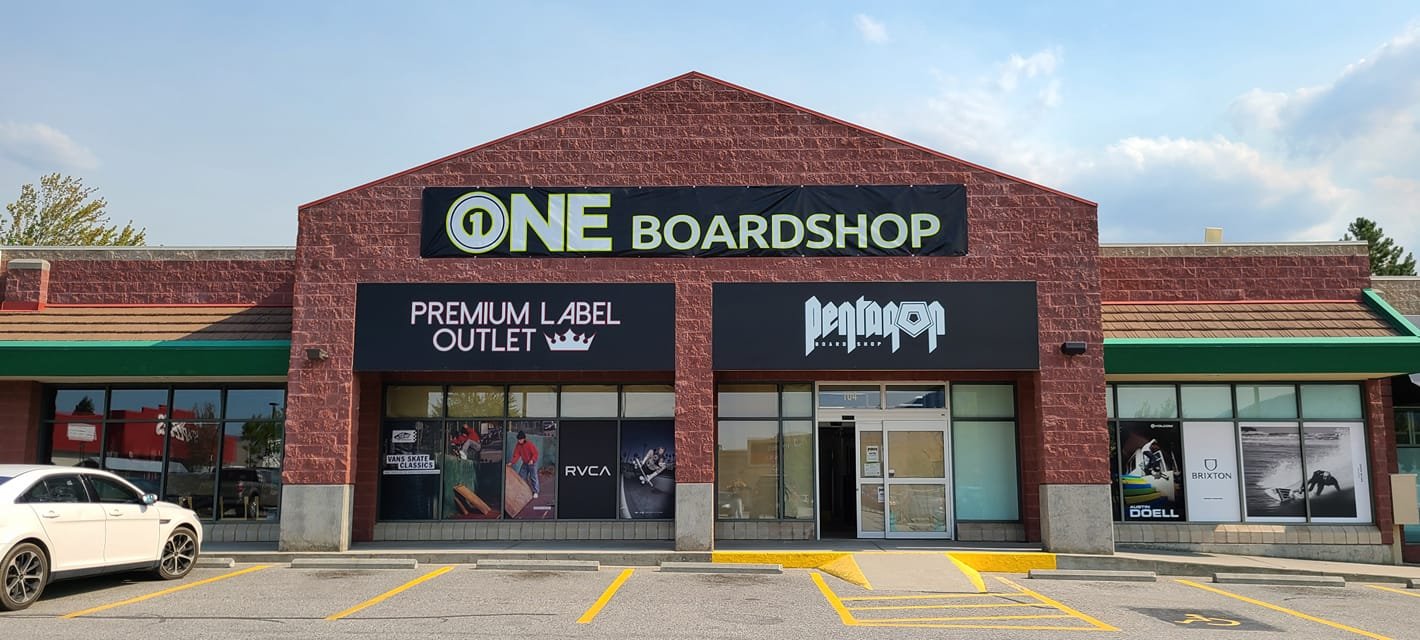 storefront of a board shop with signs for Premium Label Outlet and Pentagon. parking lot with a white car in front.