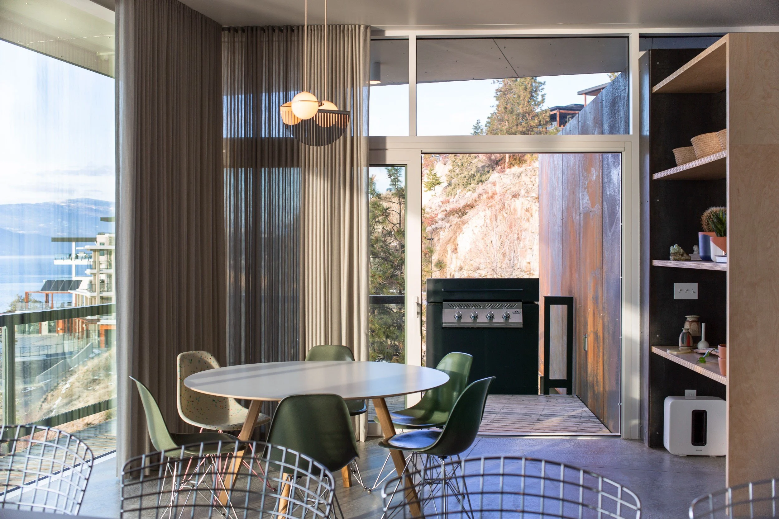 Modern indoor dining area with a round white table and green chairs, large glass sliding door leading to a balcony with a grill, mountain landscape view, and decorative items on a wooden shelf.
