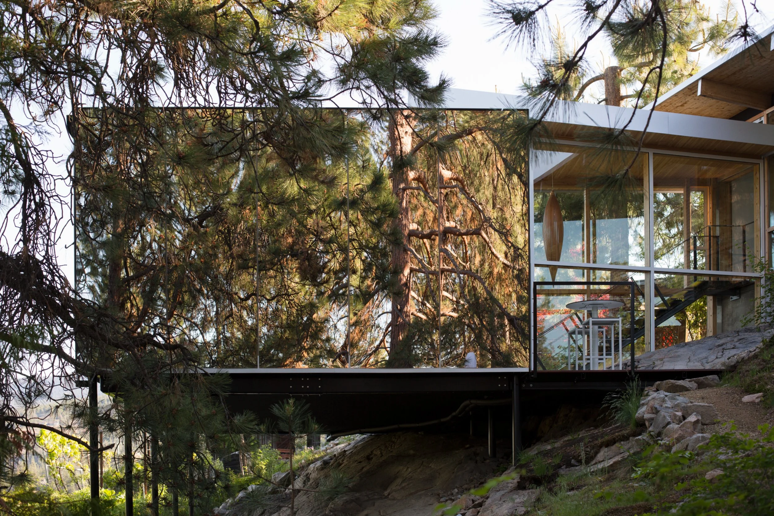 Modern house with large glass windows surrounded by trees, built on a rocky hillside with a glass railing outside.