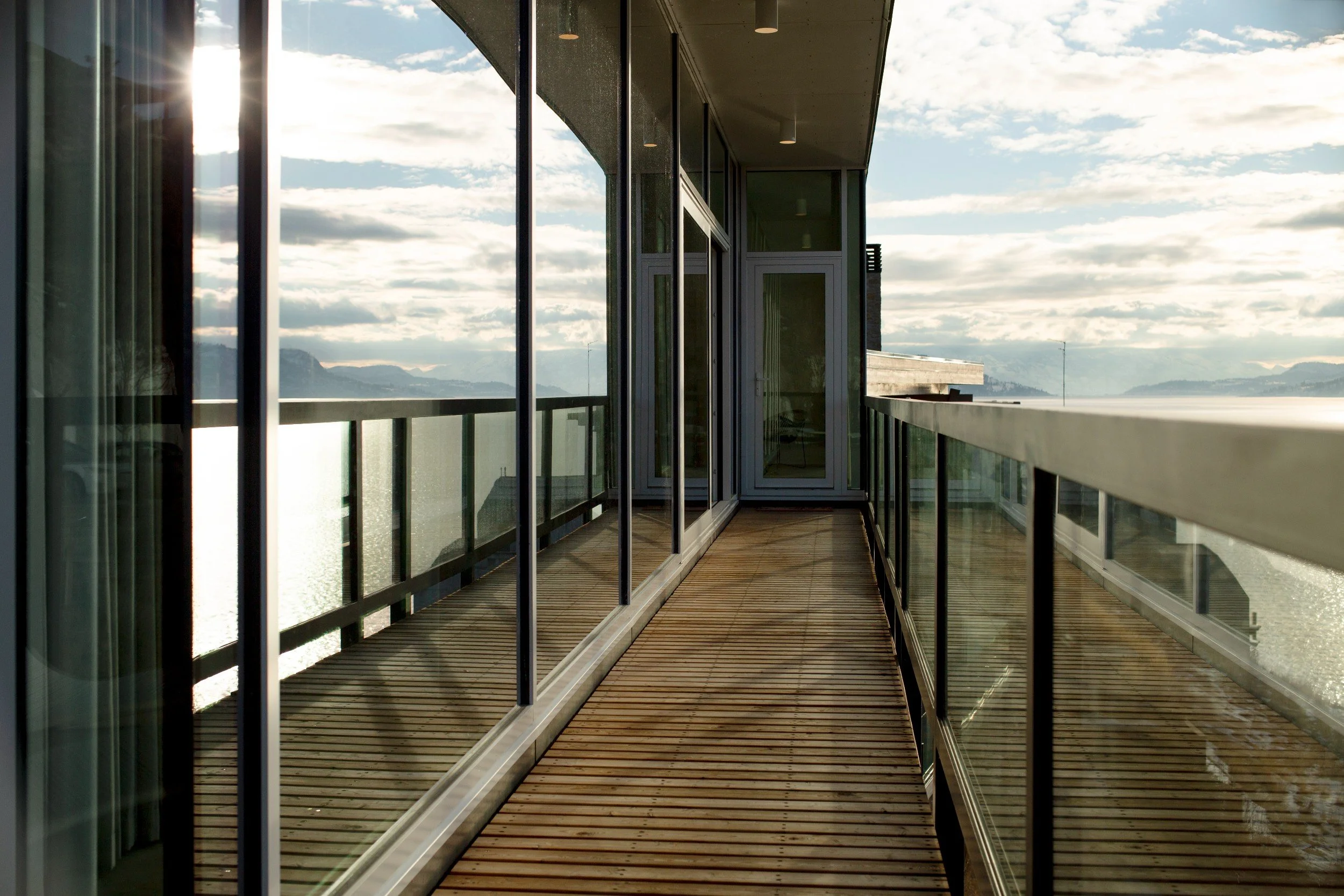 Balcony with wooden floorboards, glass railing, and sliding glass doors overlooking cloudy sky and distant mountains.