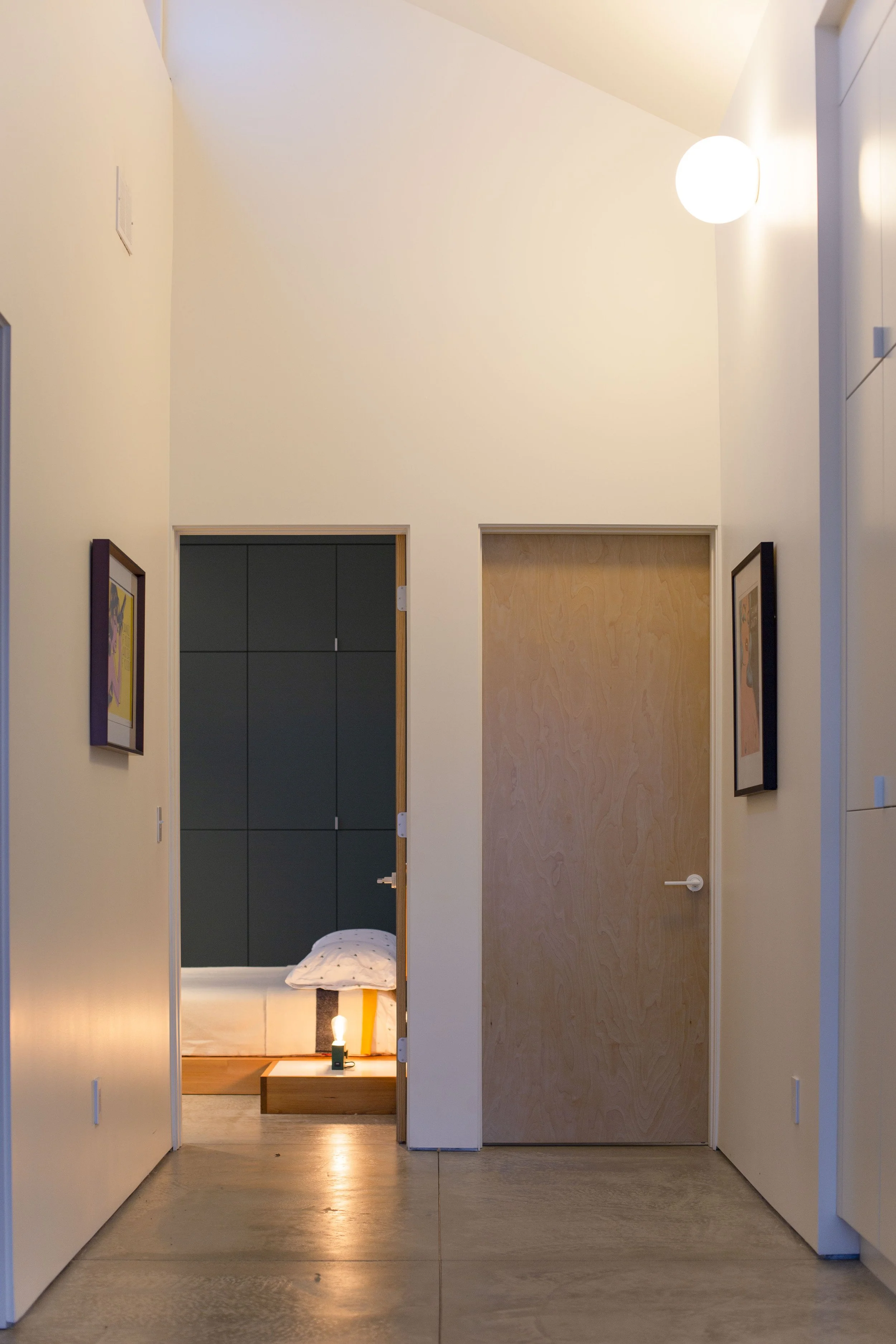A hallway with two closed wooden doors, one on each side, and a bedroom with a bed and bedside lamp visible through the open door in the middle background.