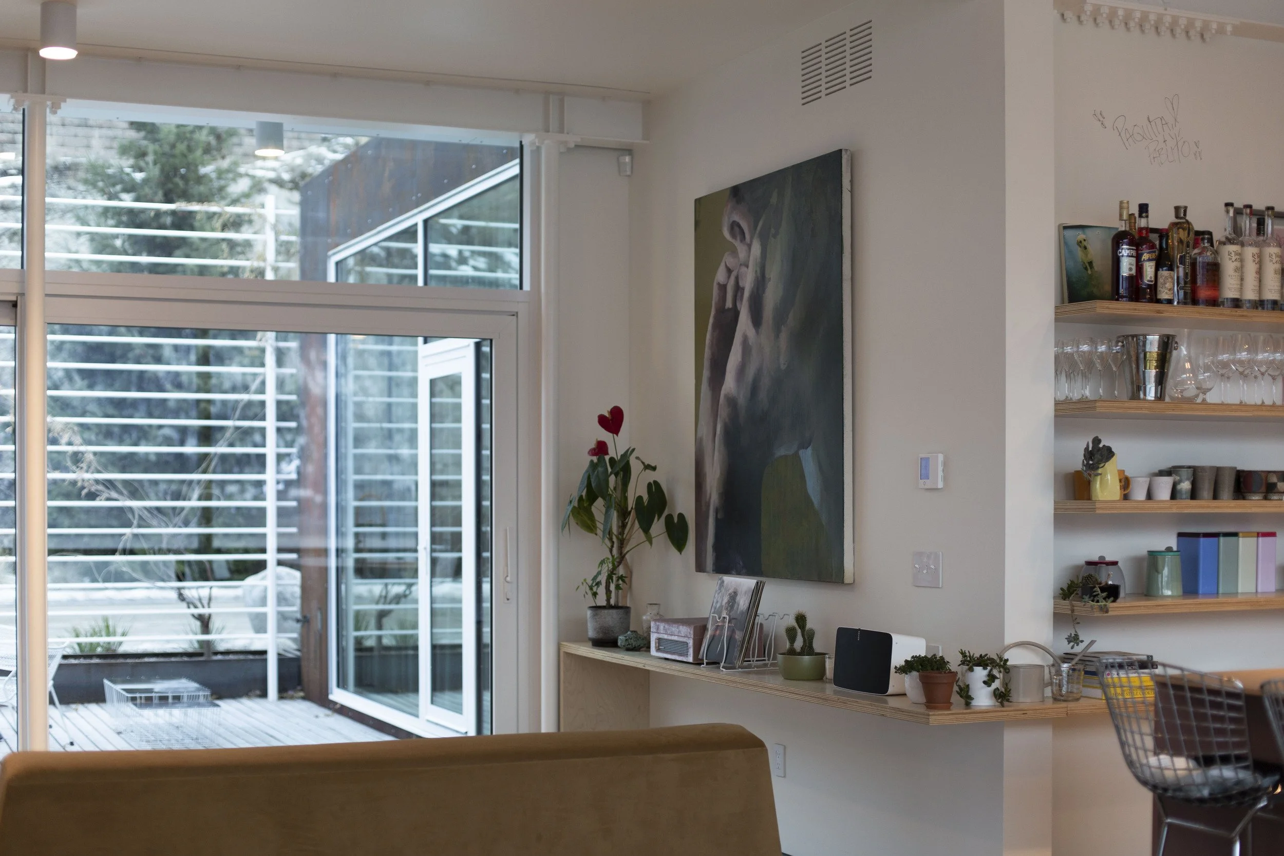 Living room with large window, an abstract portrait painting on the wall, a potted plant with heart-shaped leaves, a wooden shelf with plants and books, and a bar area with bottles and glassware.