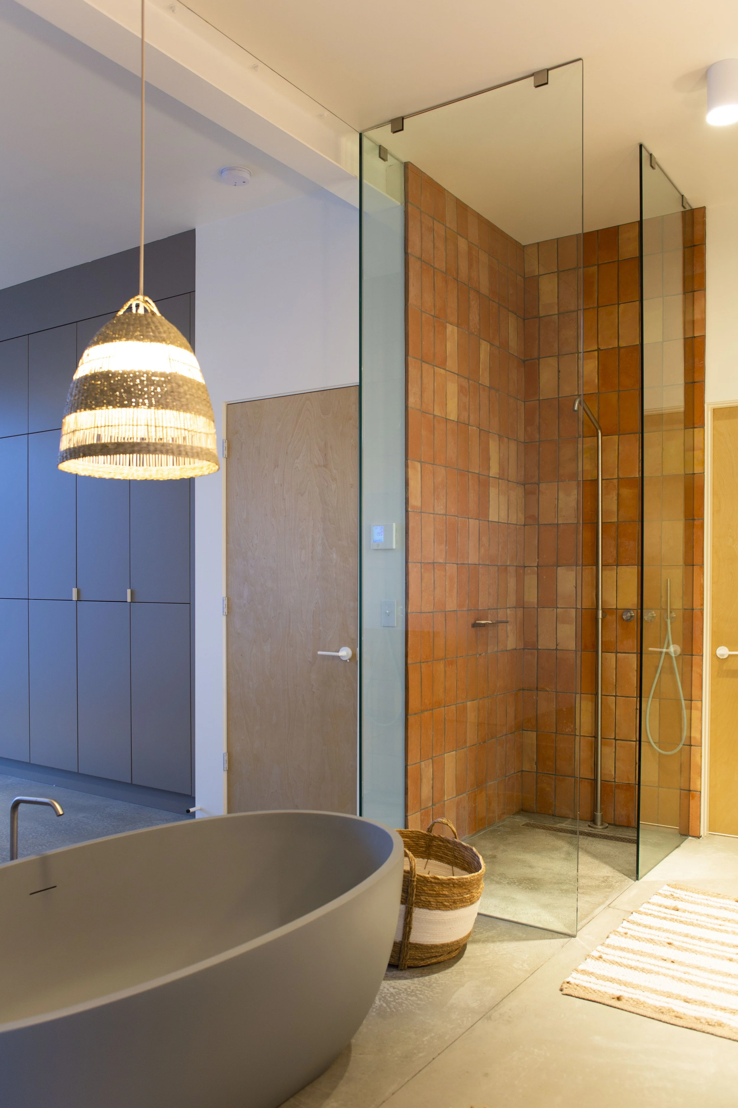Modern bathroom with a freestanding bathtub, glass-walled shower, wooden door, pendant light, cabinets, woven basket, and striped rug.