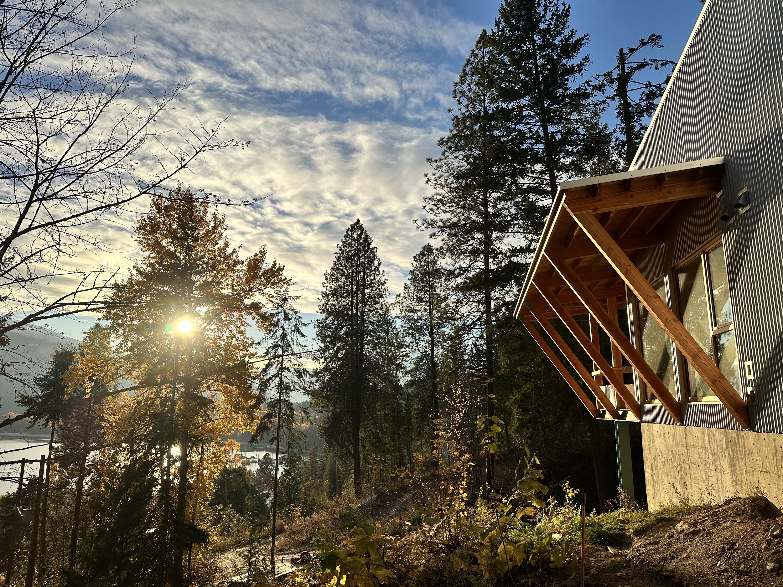 Mountain Modern house with large windows and wooden awnings situated in a wooded area with tall trees, autumn foliage, and a partly cloudy sky, illuminated by the sun near the horizon.